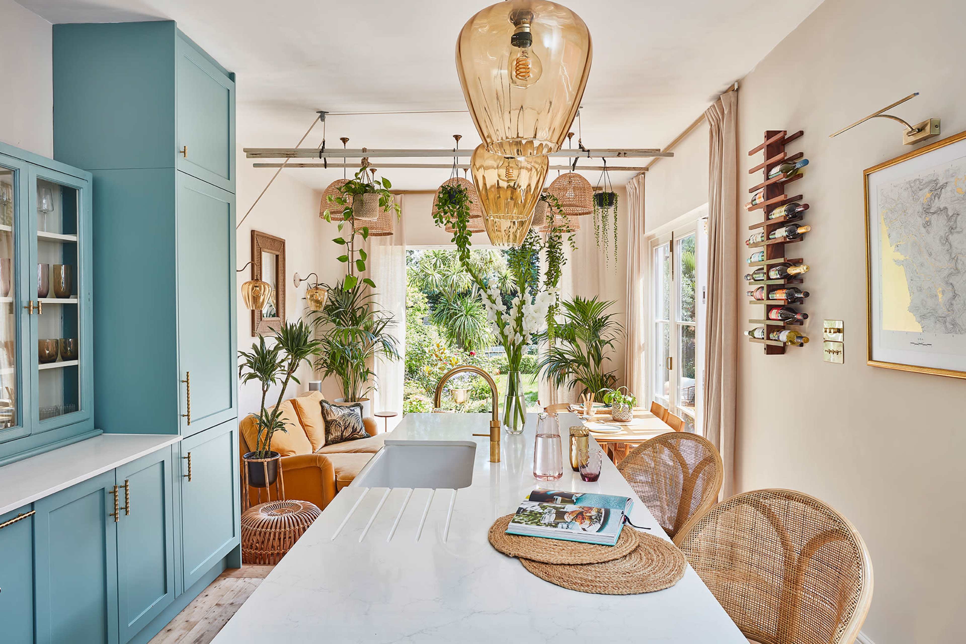 A modern kitchen with teal cabinets, a white marble countertop, and a dining area featuring wicker chairs, plants, and large windows overlooking a garden.
