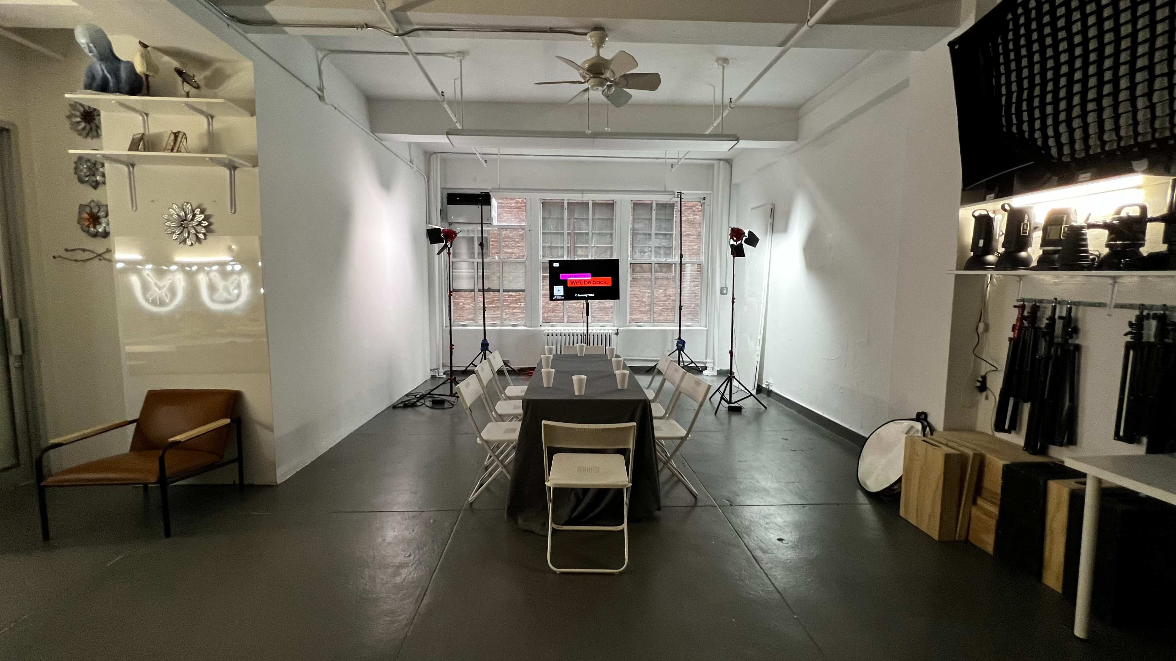 The image shows a spacious, well-lit room set up for an event, featuring a long table with chairs, a television screen at the front, and studio lighting equipment positioned around the space.