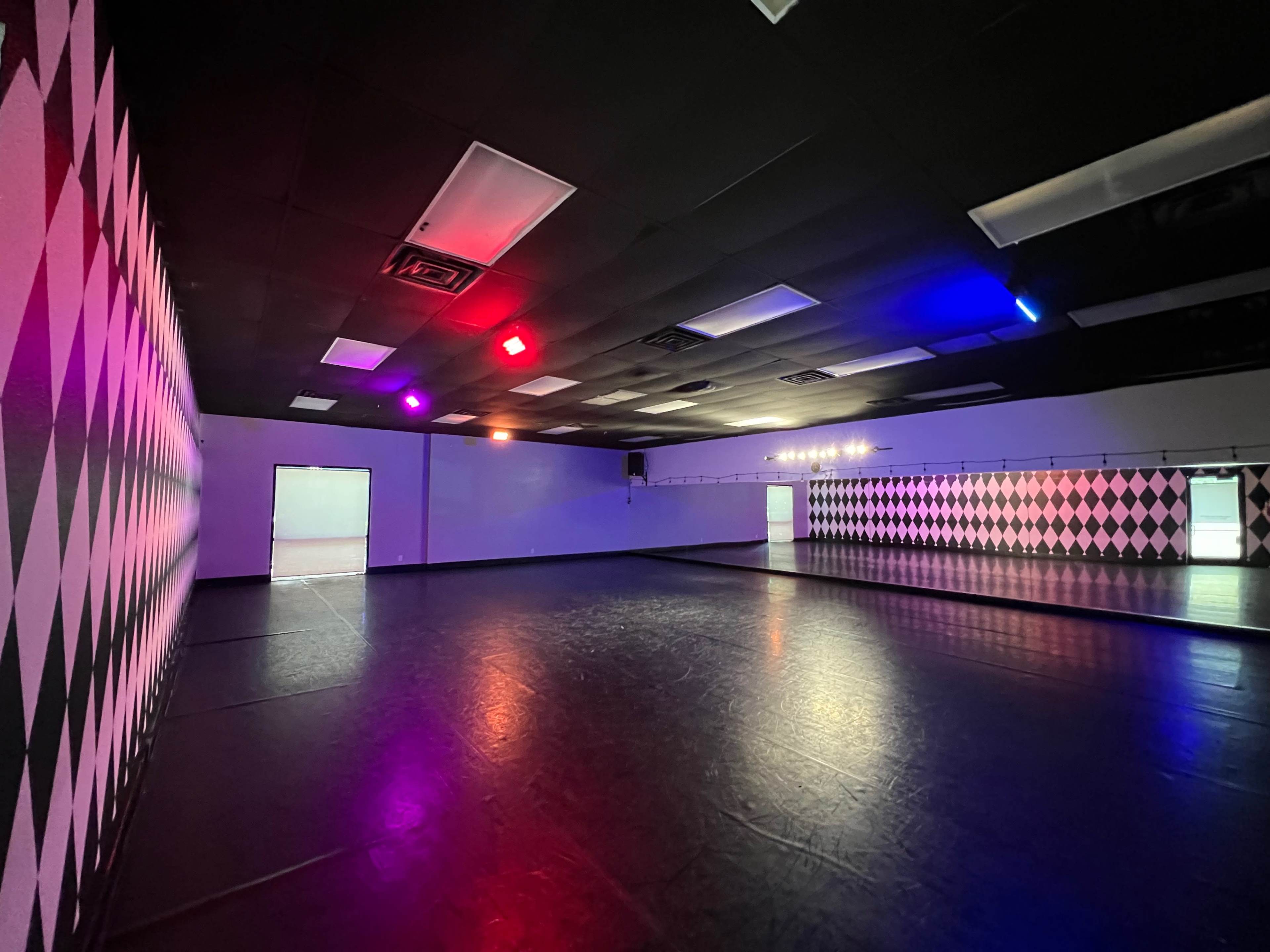 A spacious dance studio with a checkered black and white wall and colored lights illuminating the room.