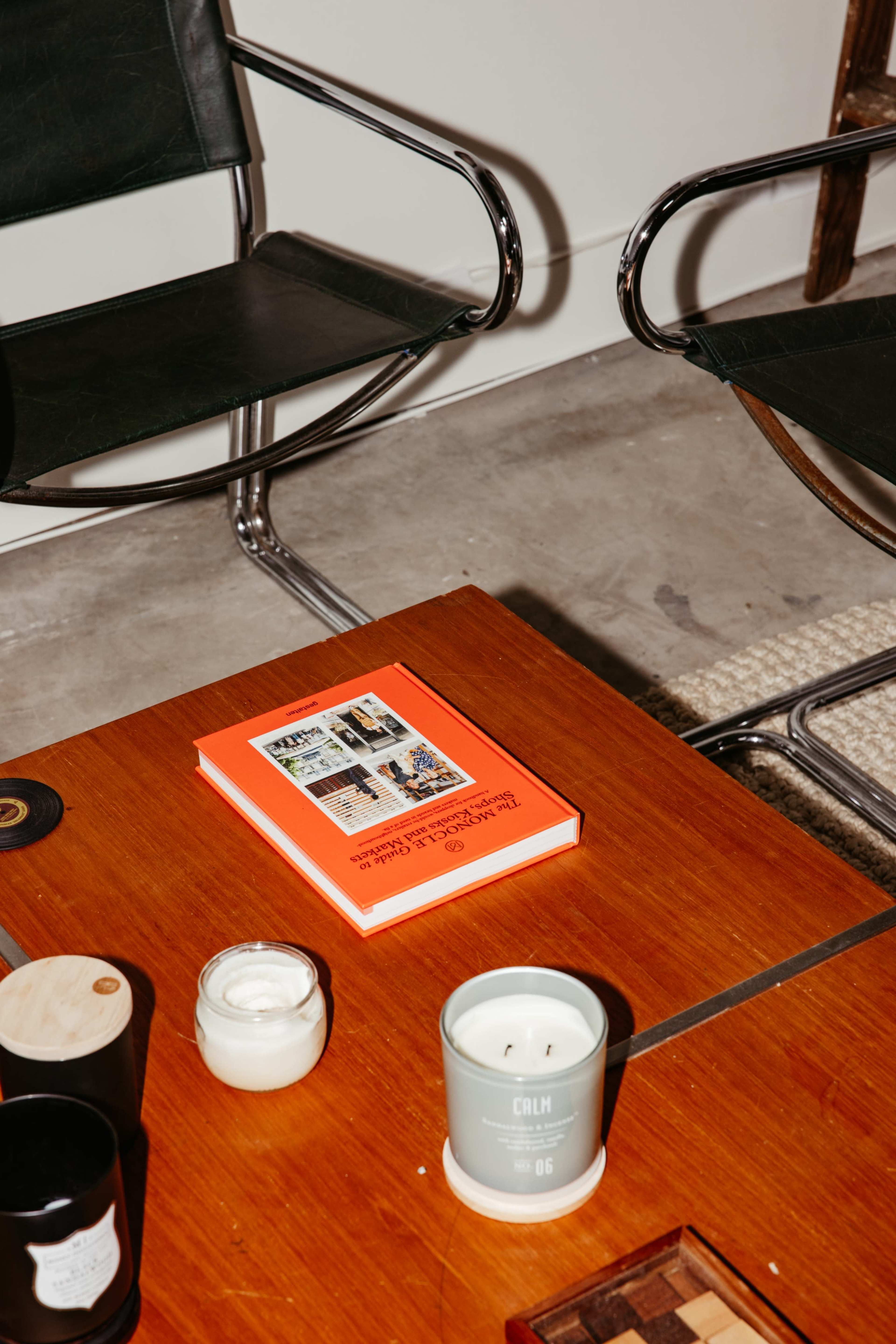 A wooden table displays an orange book, a white candle, and various small objects, while two black chairs are positioned nearby.