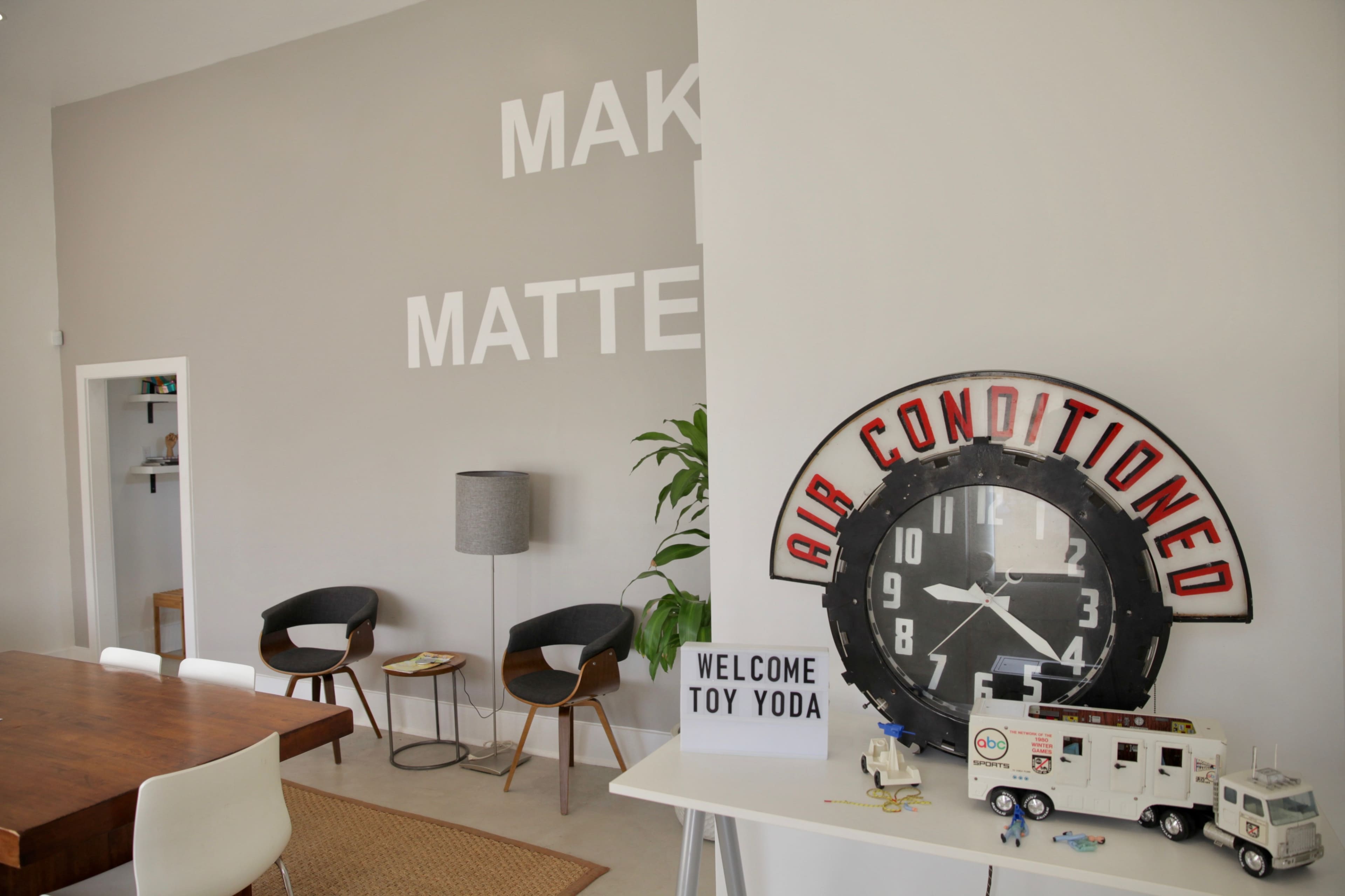 A modern room featuring a large wall with the words "MAKE IT MATTER," a wooden table, a vintage clock labeled "AIR CONDITIONED," a decorative sign reading "WELCOME TOY YODA," and minimalist furniture.