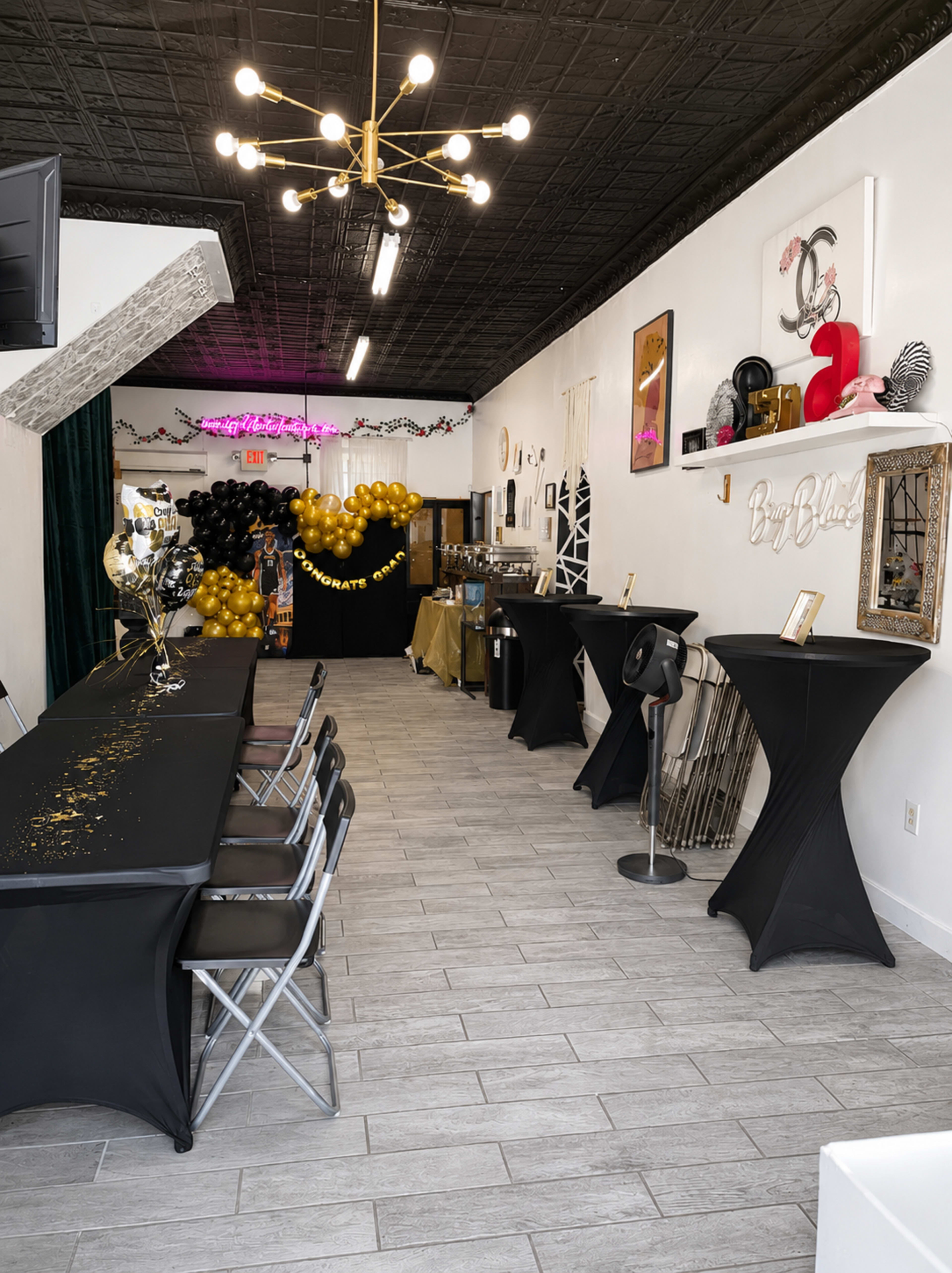 The image shows an event space decorated with black tables, gold and black balloons, and a backdrop with a heart shape, set up for a celebration.