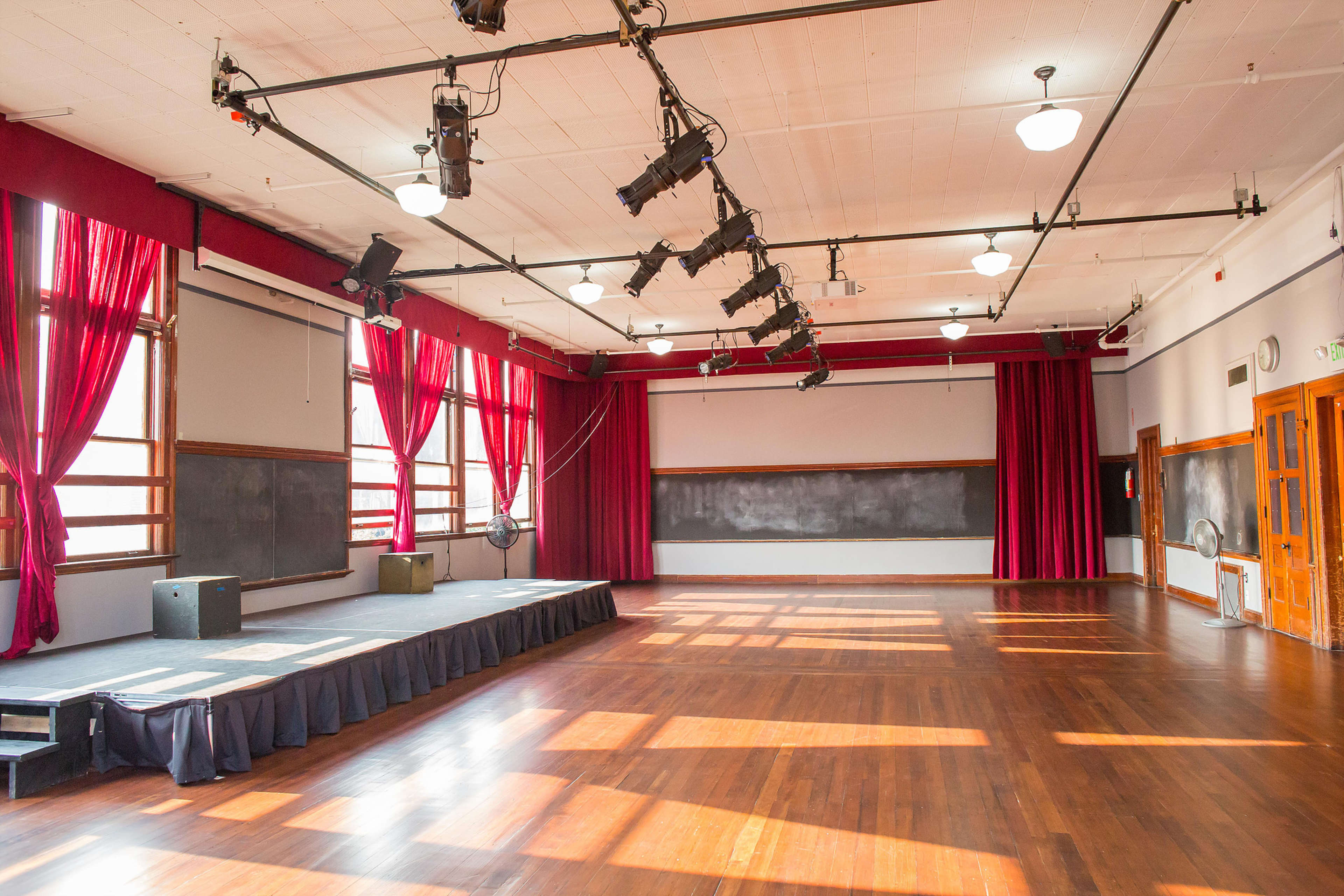 A spacious room with large windows, wooden floors, red curtains, and a stage equipped with spotlights and sound equipment.