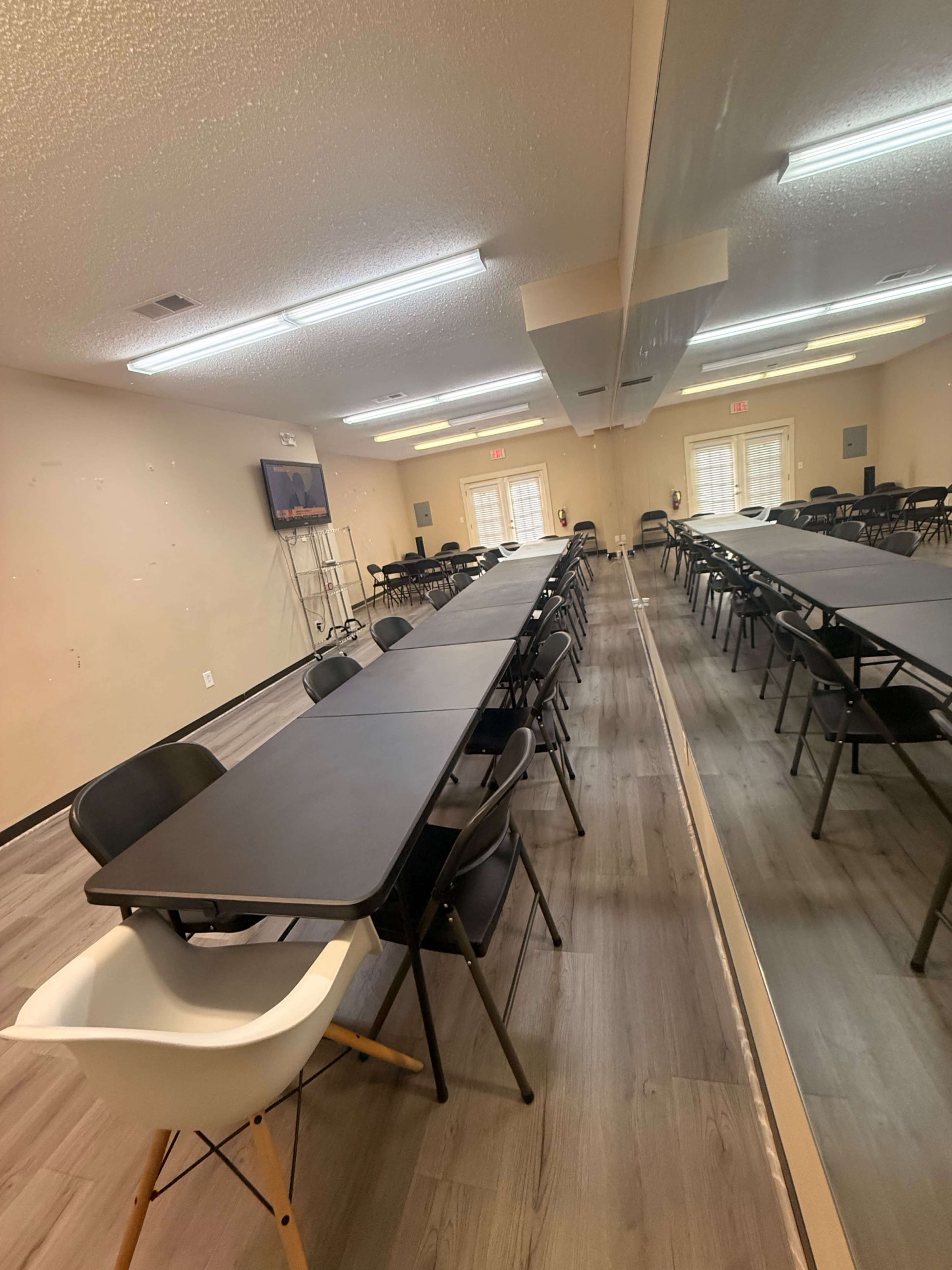 The image shows a spacious room set up with multiple rows of black folding tables and chairs, featuring a large mirror on one wall and a television screen mounted on another.