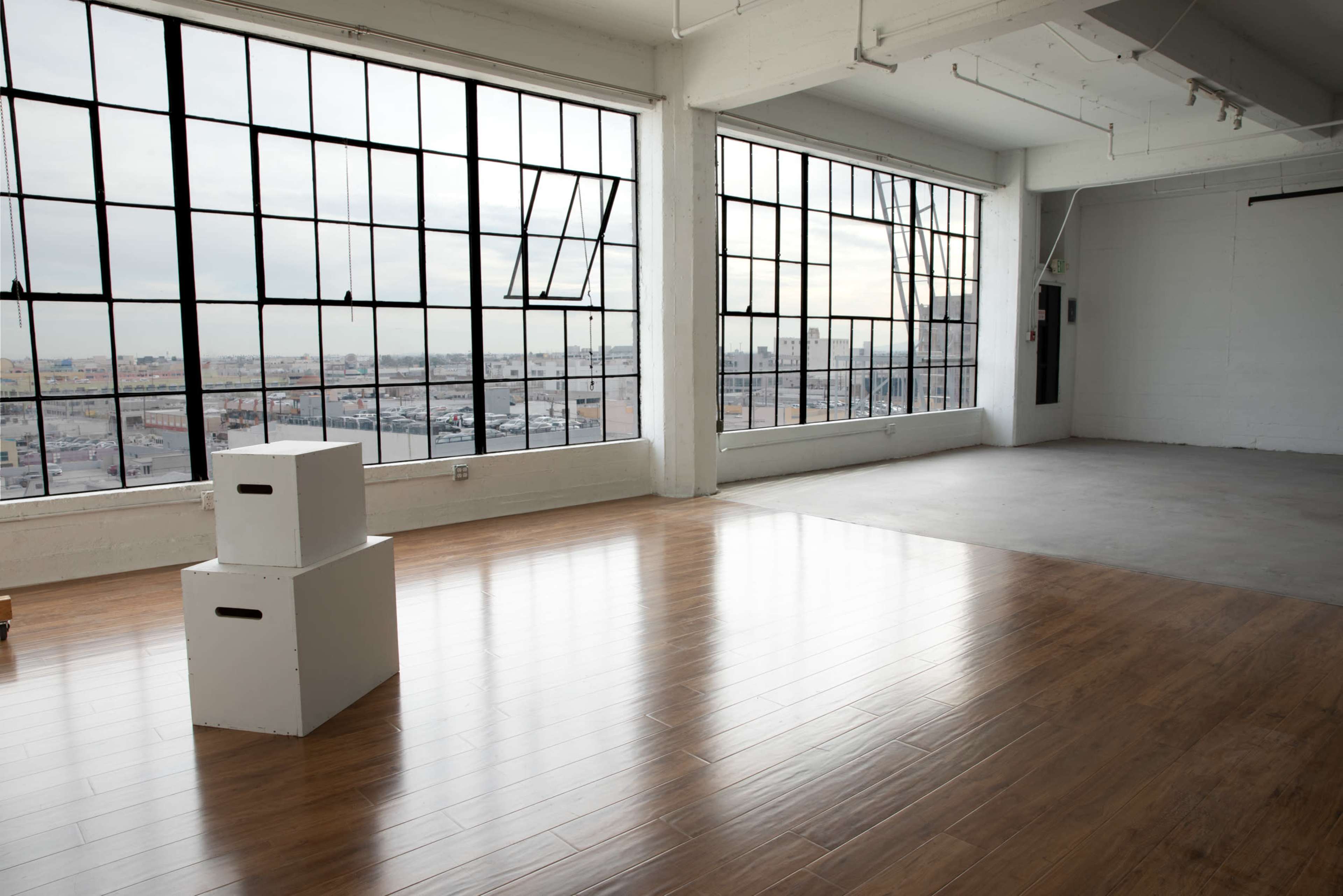 The image shows a spacious, empty room with large windows, wooden flooring, and two white cardboard boxes on the floor.