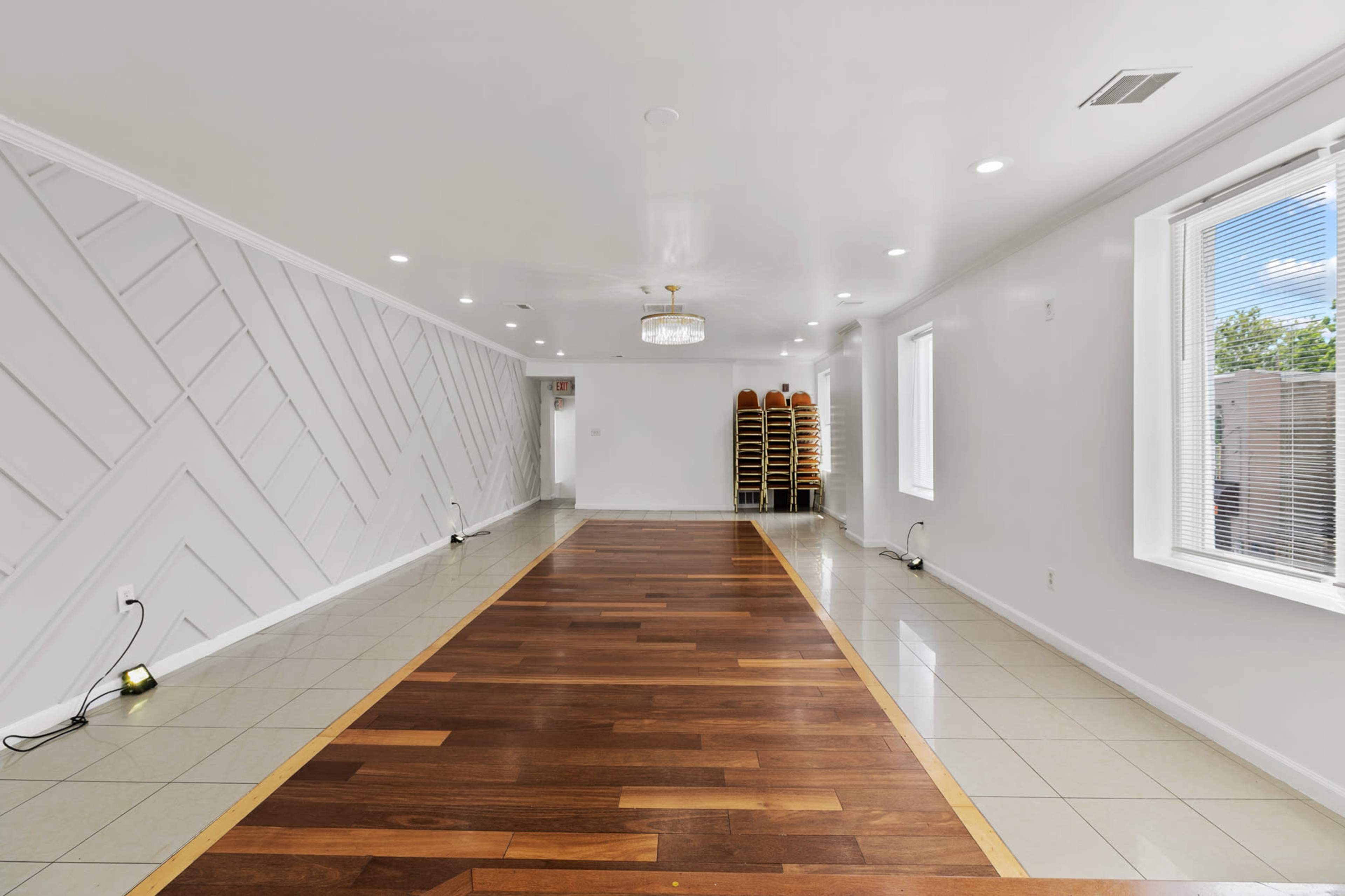 A spacious, empty room with a combination of tiled and wooden flooring, featuring a decorative wall and stacks of chairs against one wall.
