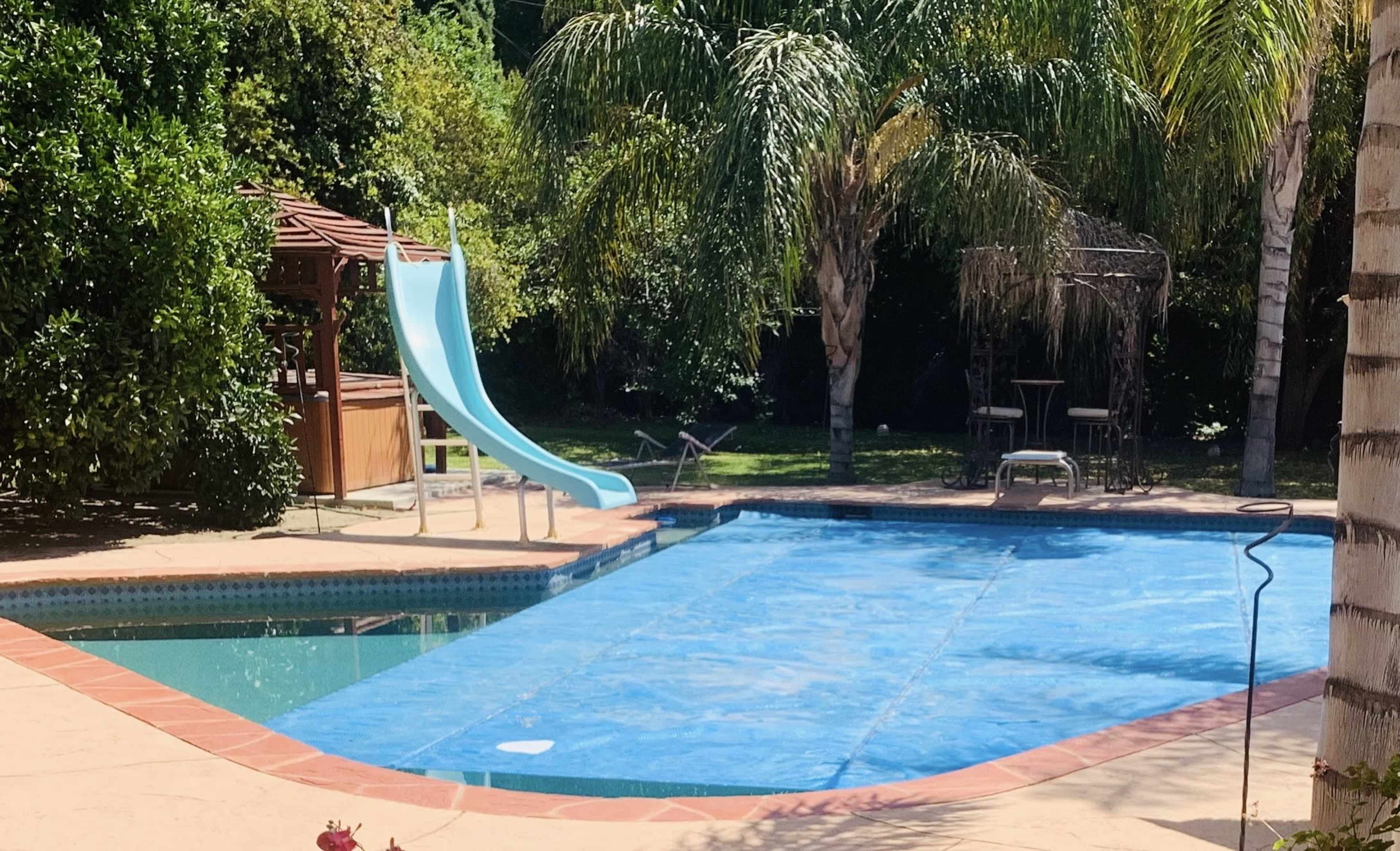 A backyard swimming pool is covered with a blue tarp, featuring a slide and surrounded by palm trees and outdoor furniture.