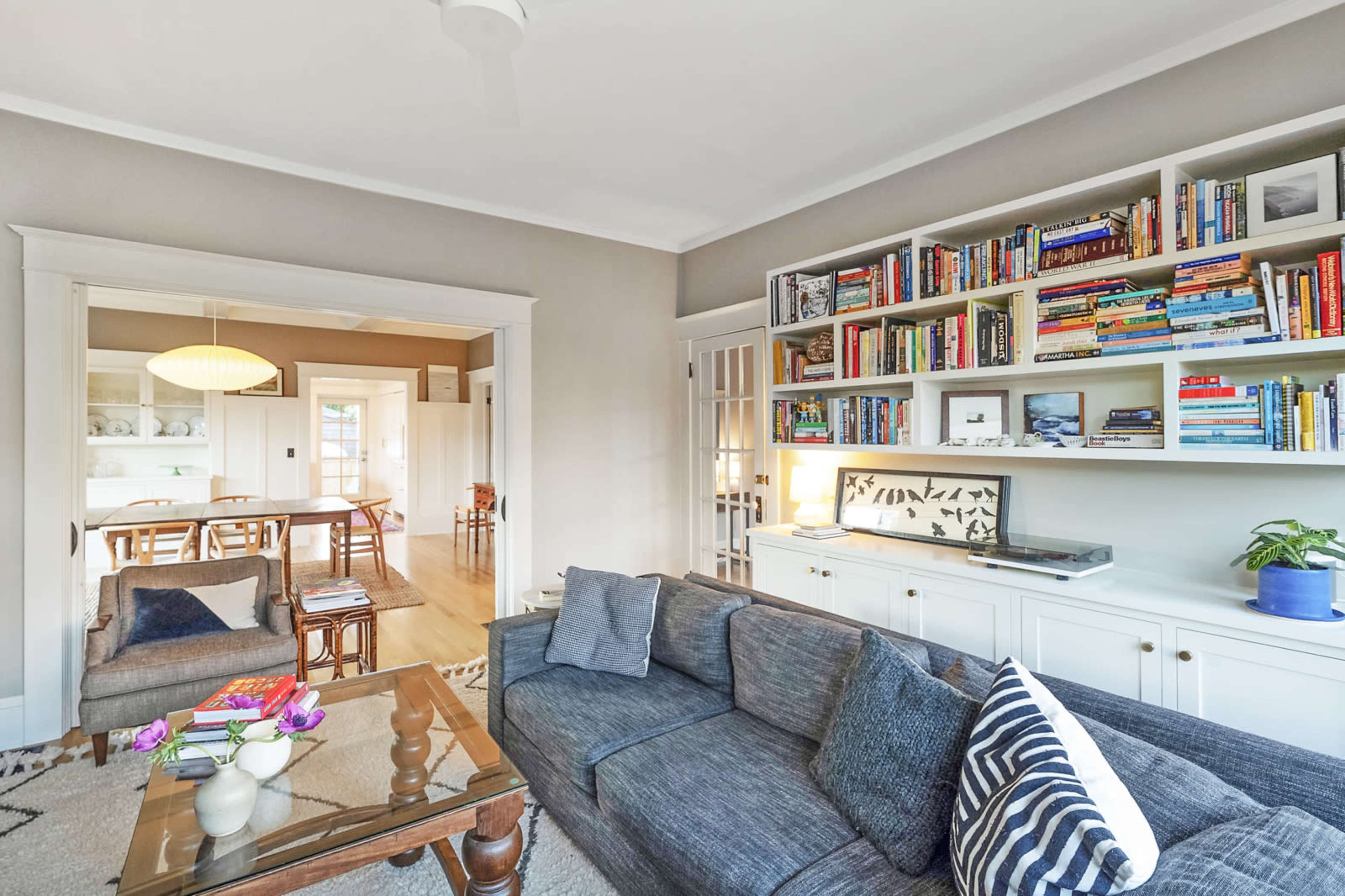 A living room with a gray sofa, a glass coffee table, and a bookshelf filled with books, connected to a dining area visible in the background.