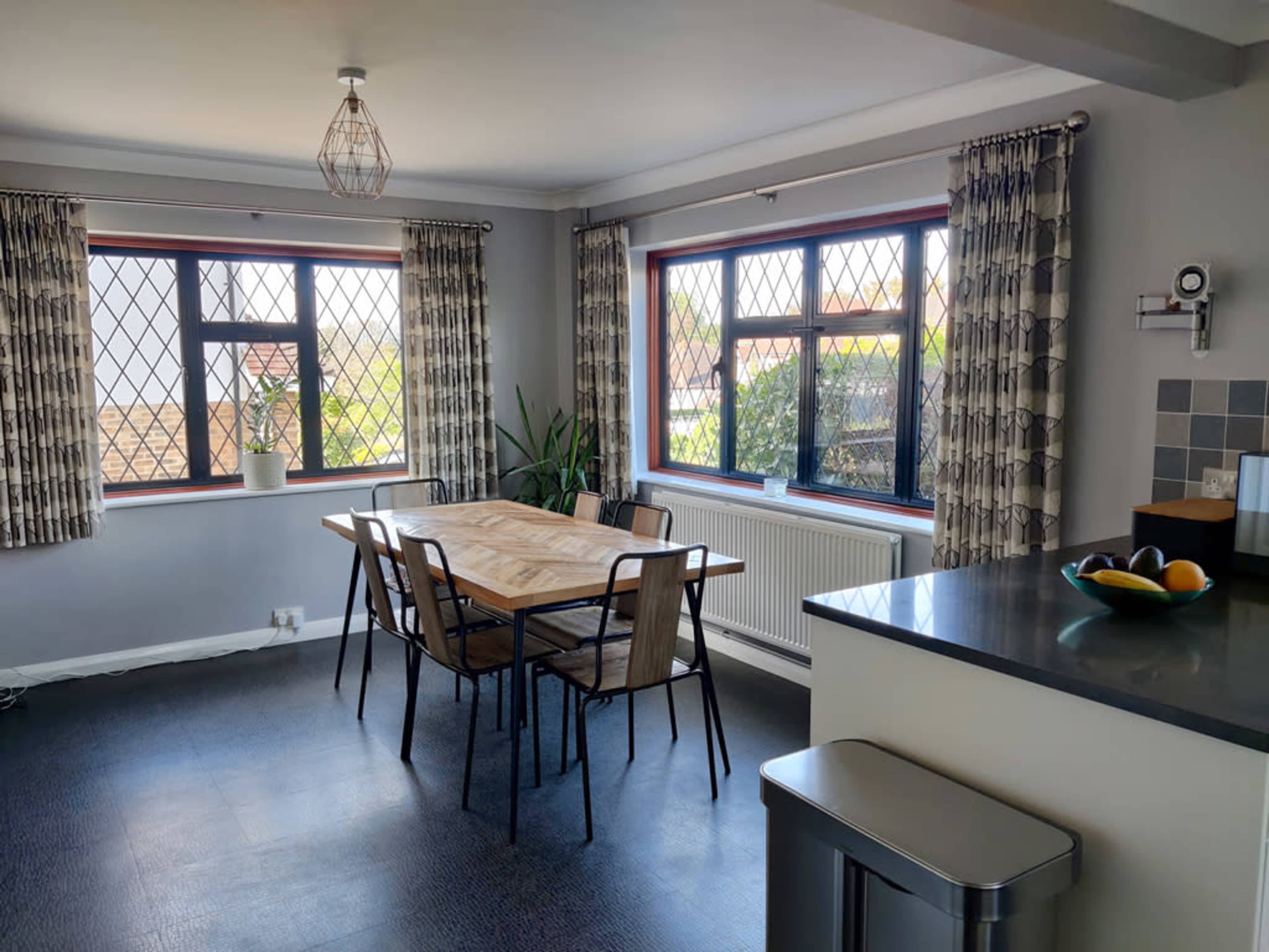A dining area features a wooden table surrounded by metal chairs, with large windows allowing natural light and greenery visible outside.