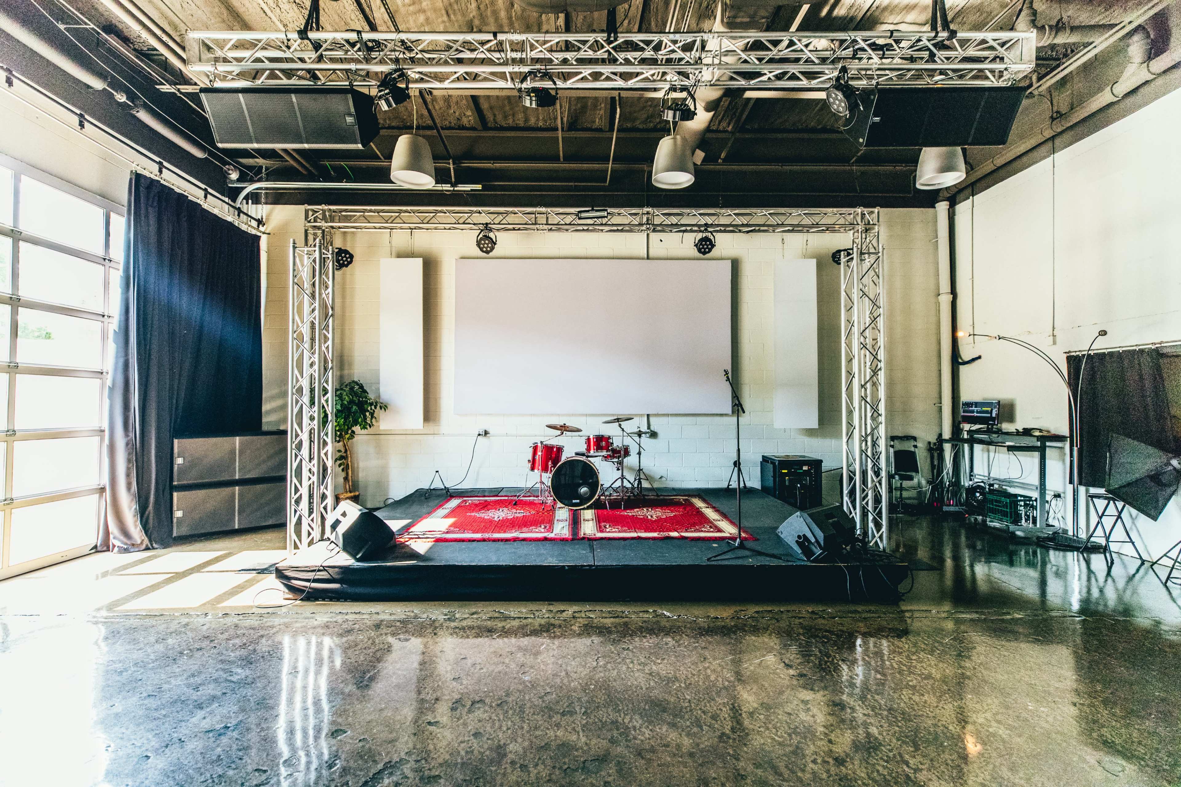 A performance stage with a drum set, sound equipment, and a large screen is set up in a room with a polished concrete floor and industrial lighting.