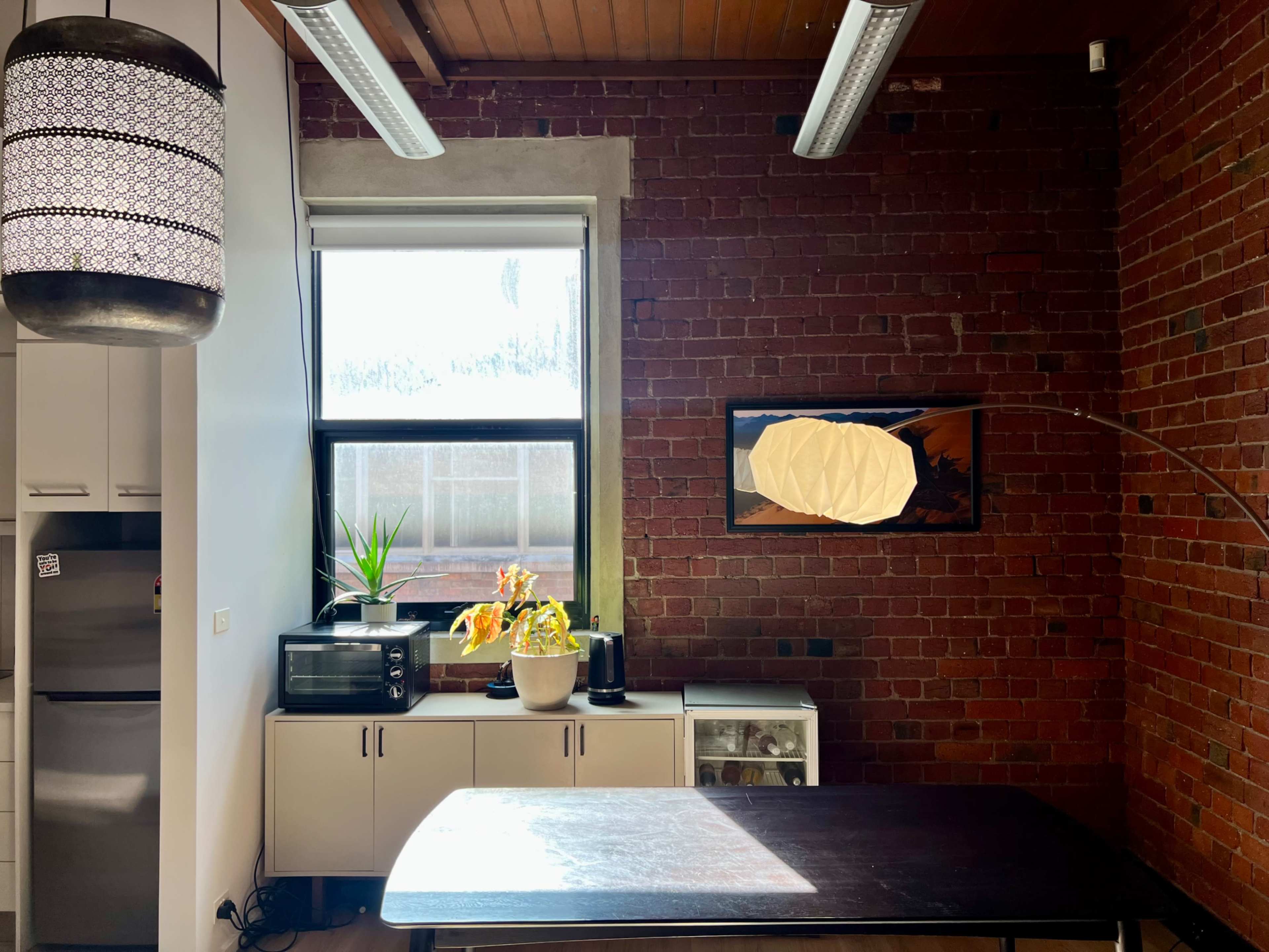 The image showcases a modern kitchen with exposed brick walls, a large window, an indoor plant, and a dining table in the center.