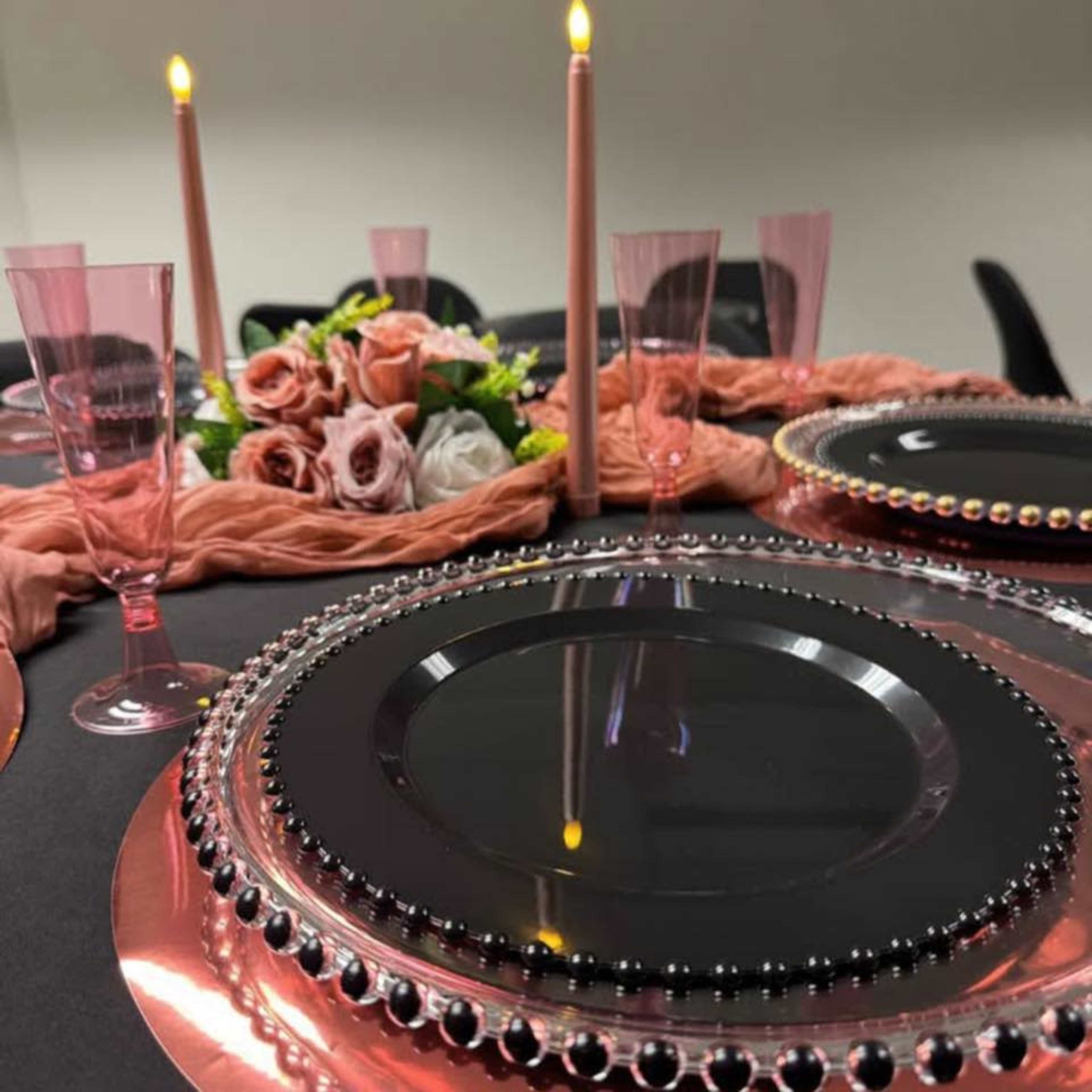 The image shows a dining table set with black plates, pink glassware, candles, and a floral centerpiece.