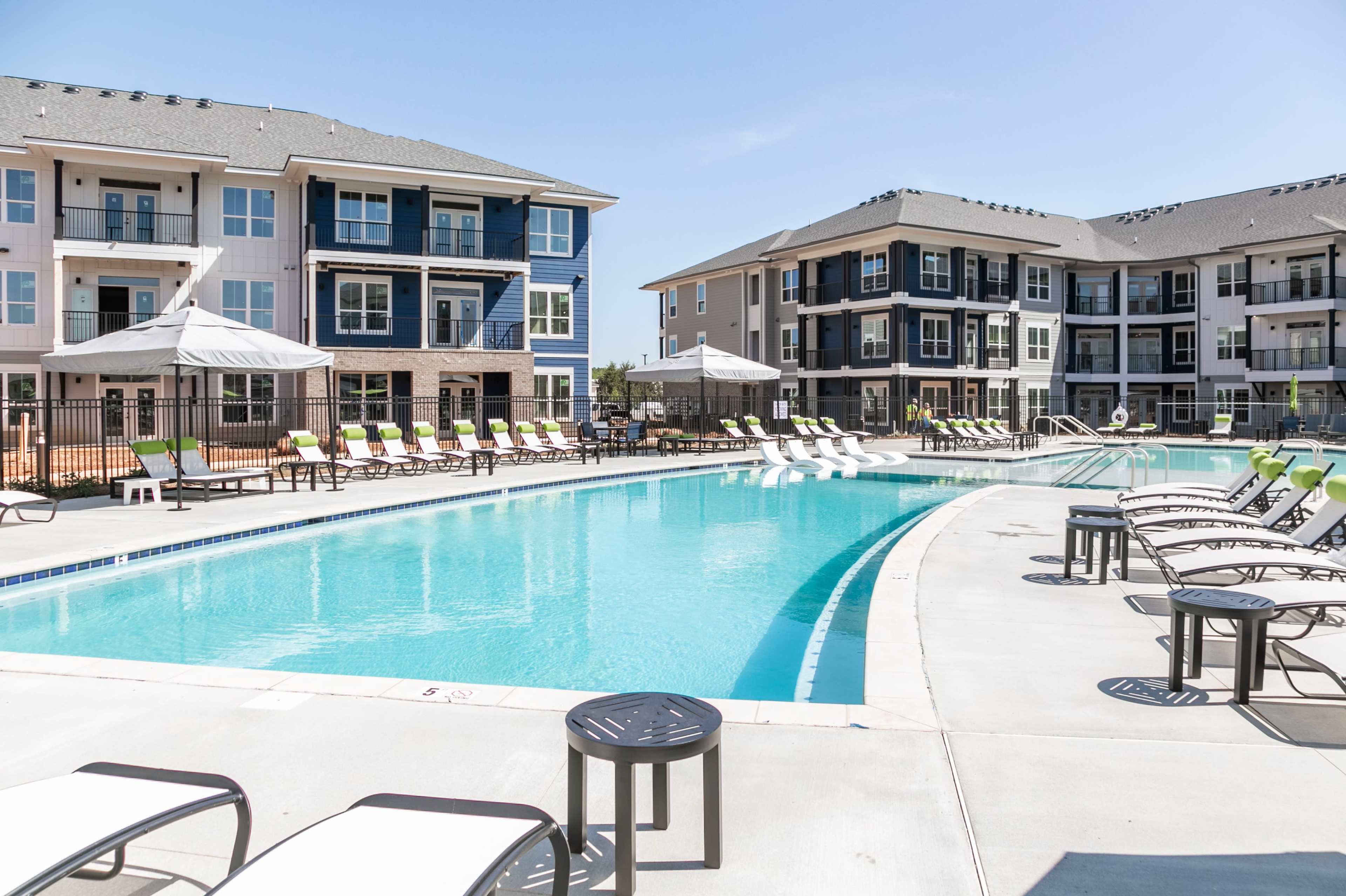 The image shows a swimming pool surrounded by lounge chairs and two multi-story residential buildings in a modern complex.