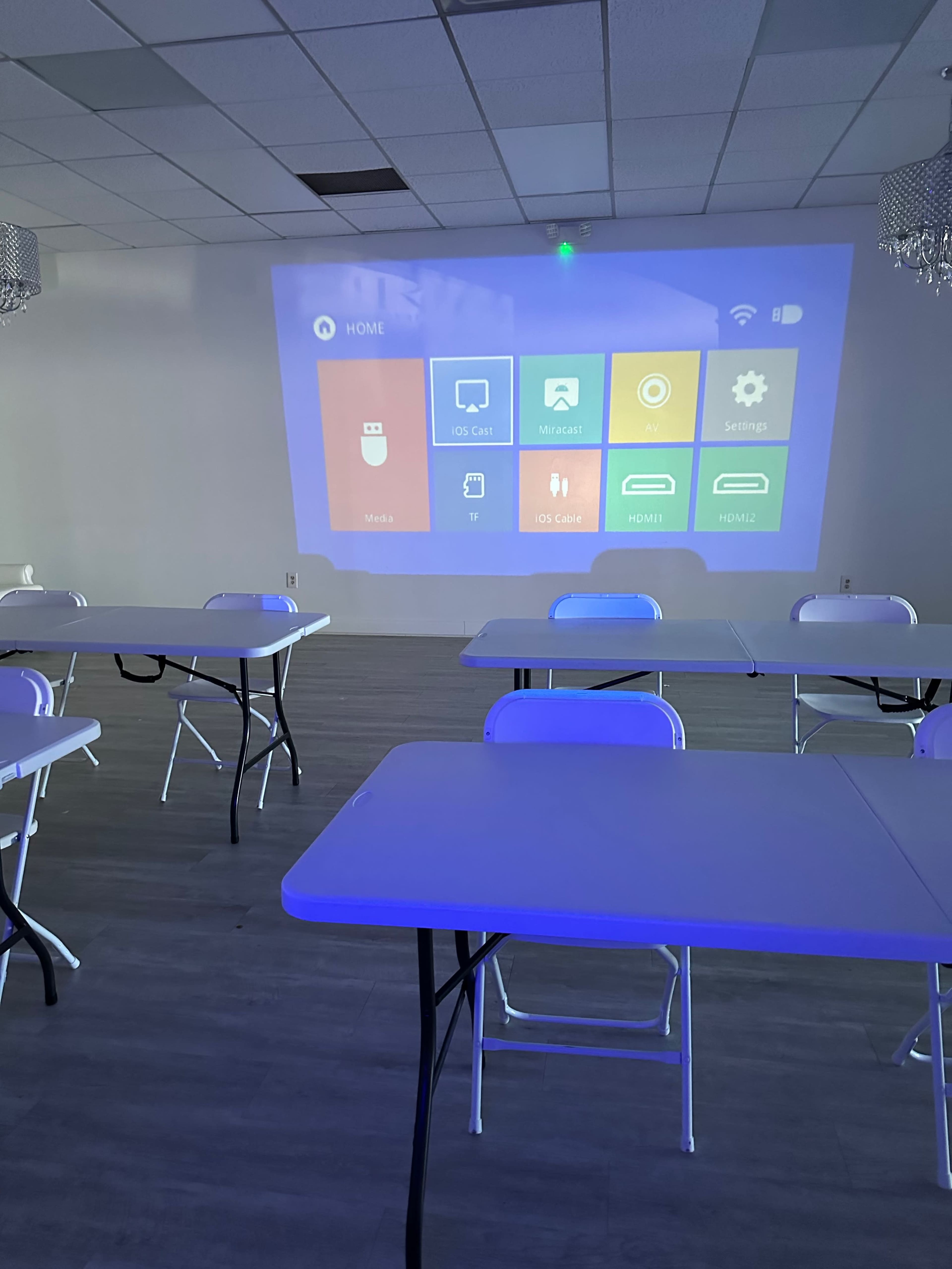 A spacious room with white tables and chairs facing a wall where a projector displays a user interface with various digital options.