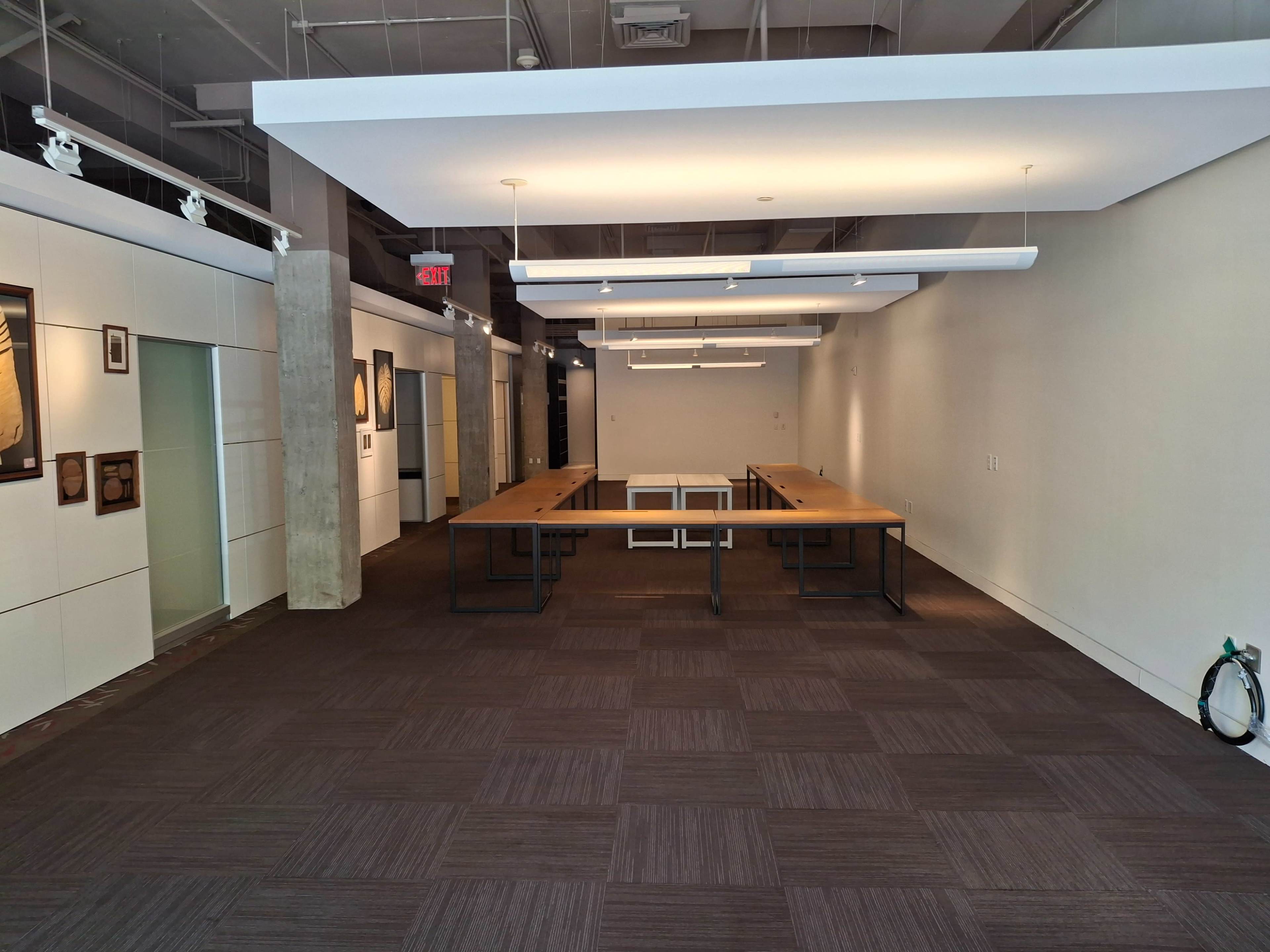 The image shows a modern office space with large wooden tables arranged in the center and gray carpet flooring.