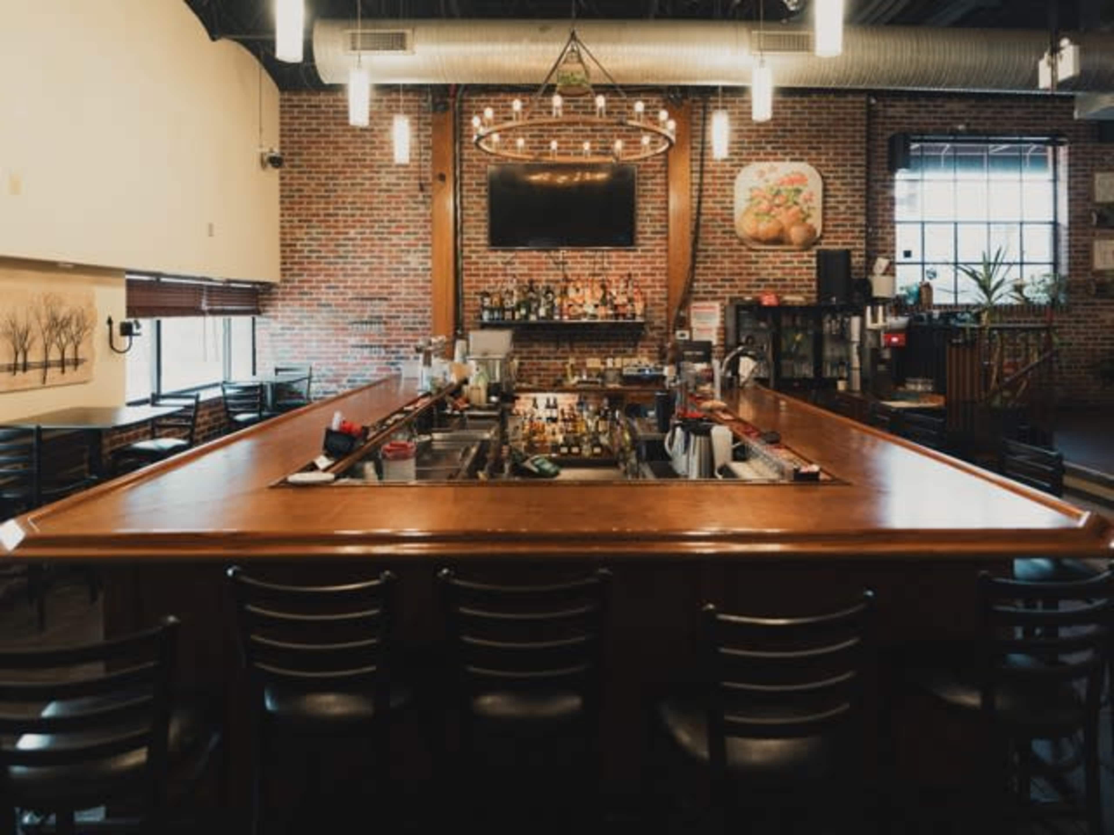 A large wooden bar with several empty black chairs surrounds it, set against a backdrop of exposed brick walls and a television mounted above a well-stocked shelf of bottles.