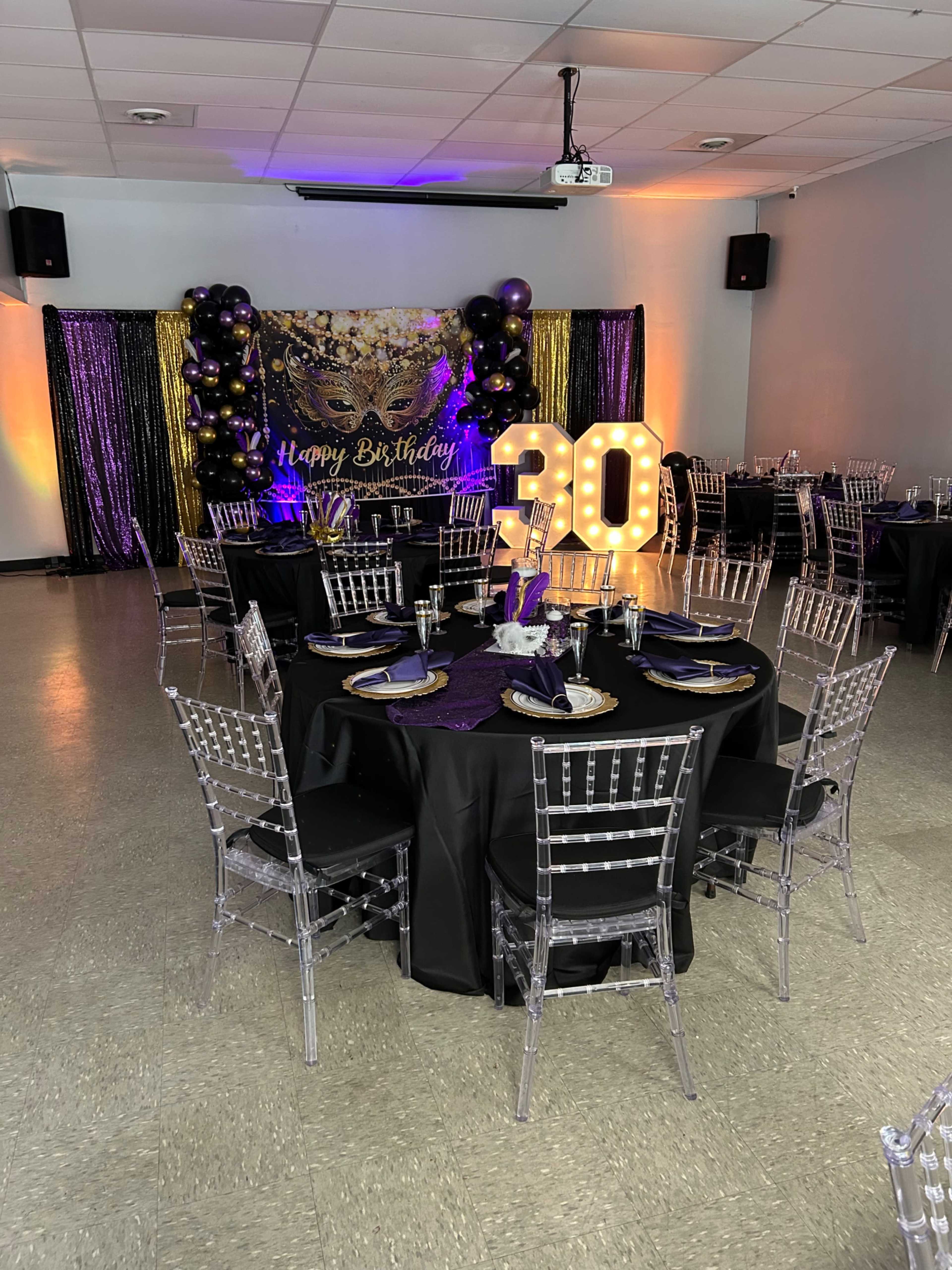 The image shows a birthday party setup with elegantly decorated tables, featuring black tablecloths, clear chairs, and a vibrant balloon arch with a "Happy Birthday" sign and a large "30" centerpiece.