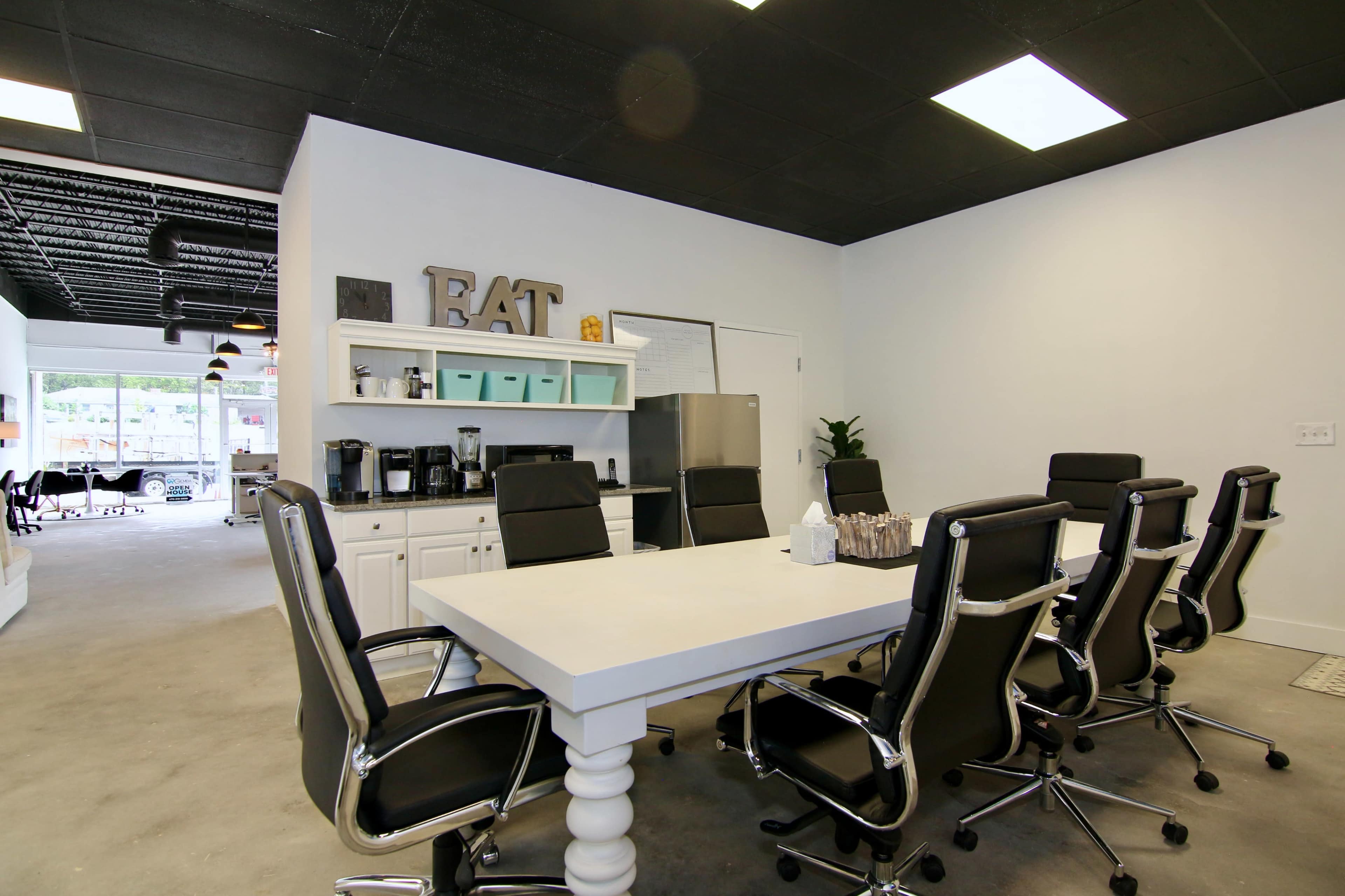 A modern conference room features a large white table surrounded by black rolling chairs, with a kitchenette area visible in the background.