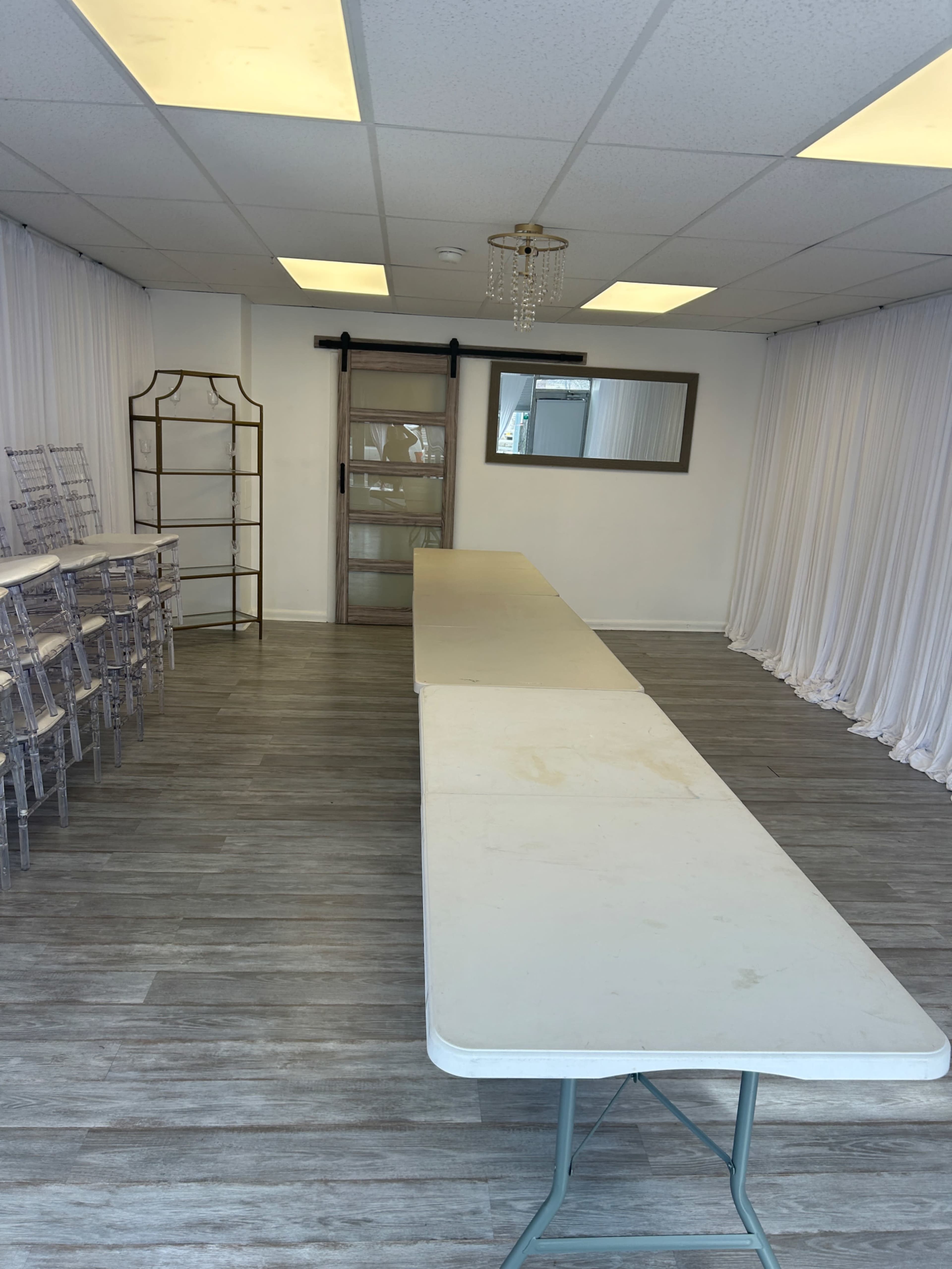 The image shows a spacious room with a long, rectangular table, a row of clear chairs stacked against the wall, and white drapes hanging from the ceiling.