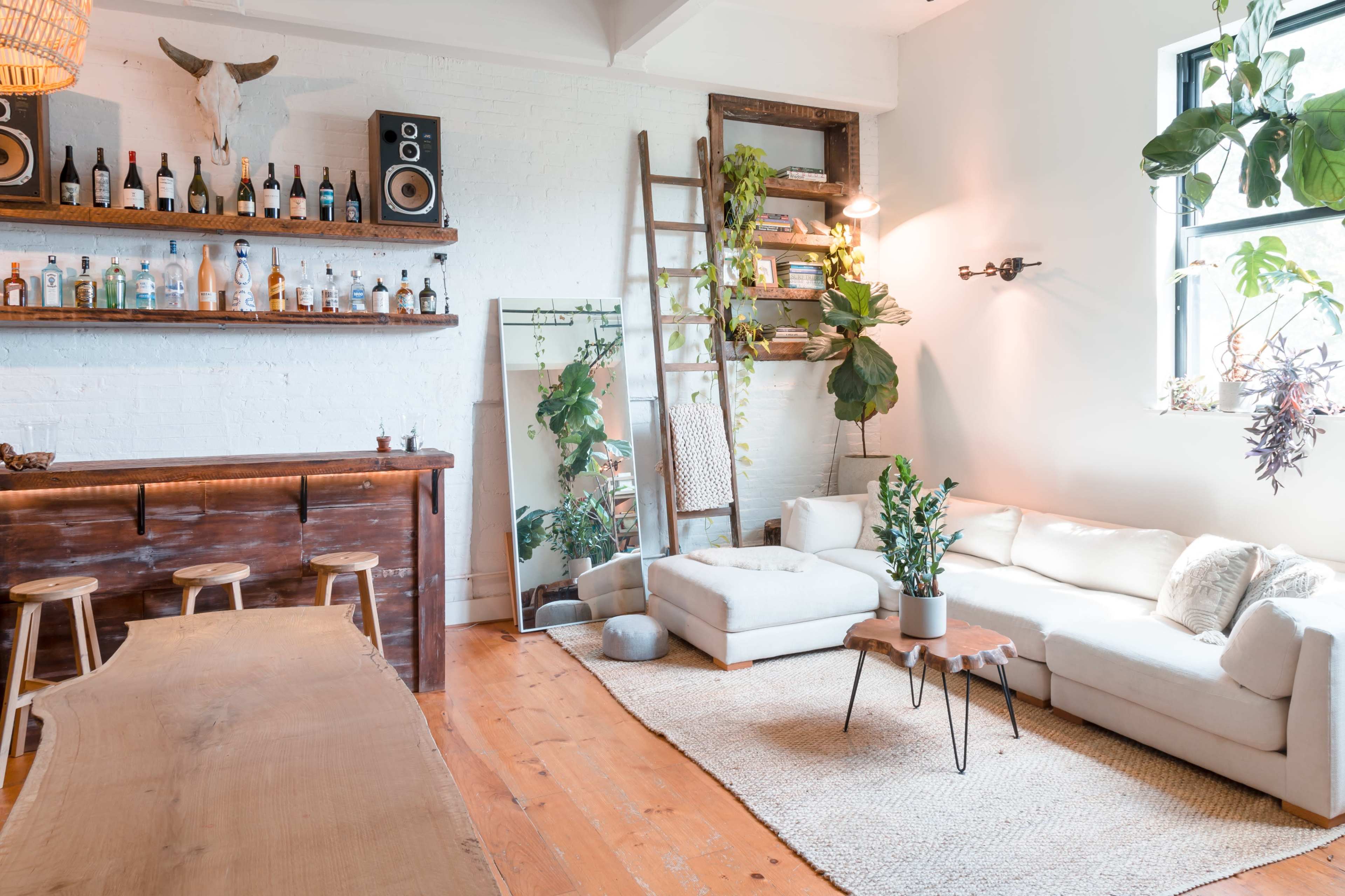 A bright, minimalist living space features a wooden bar with bottles, a ladder for plant display, a large mirror, and a light-colored sectional sofa with a low coffee table.