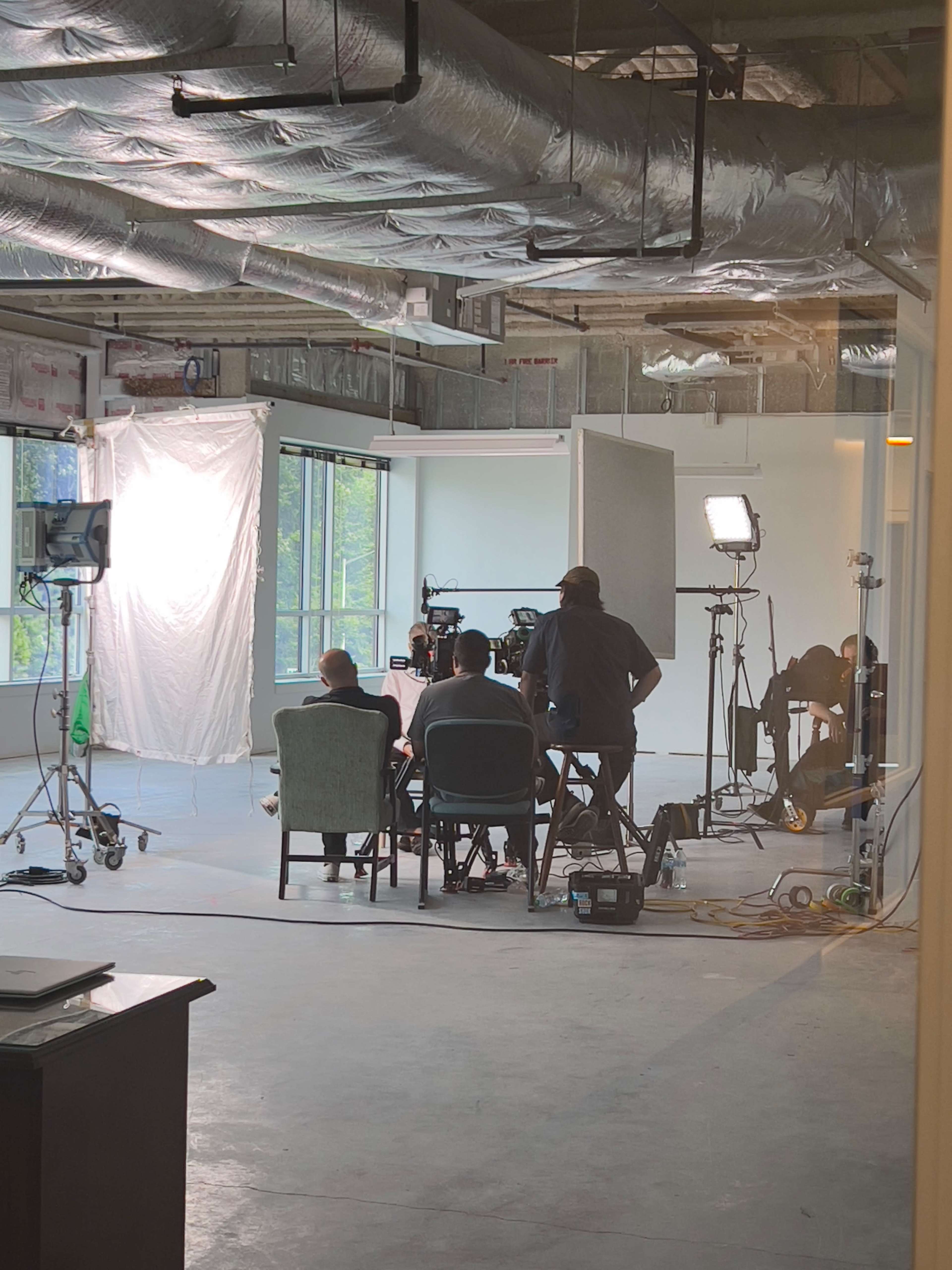 A group of people is filmed in a well-lit room with a large window, surrounded by studio equipment.
