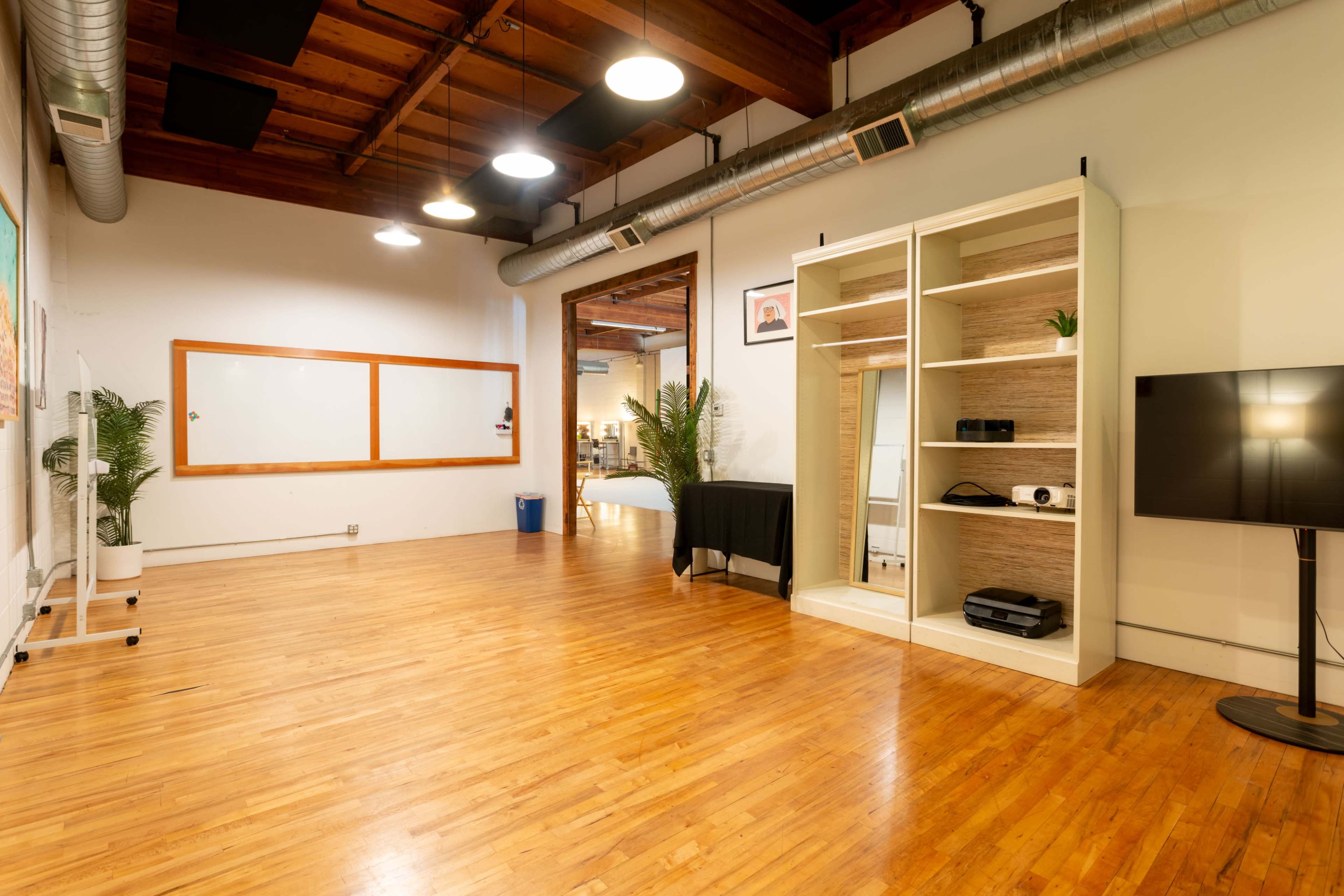 A spacious, well-lit room with wooden floors, featuring a whiteboard, a black table, and a shelving unit with equipment.