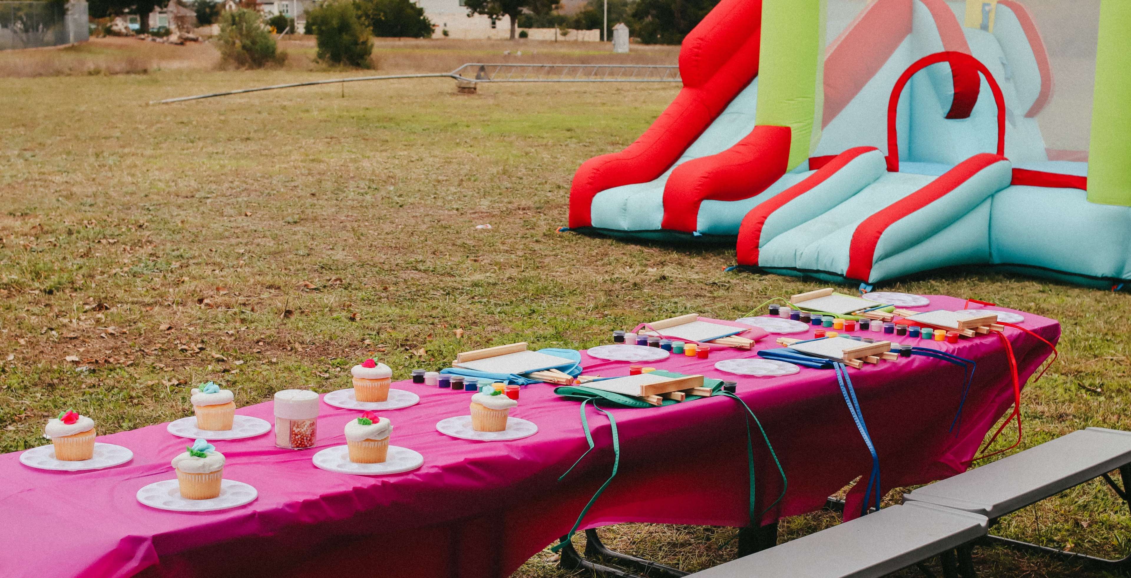 A table is set with cupcakes and colorful party supplies next to a brightly colored inflatable slide in a grassy area.