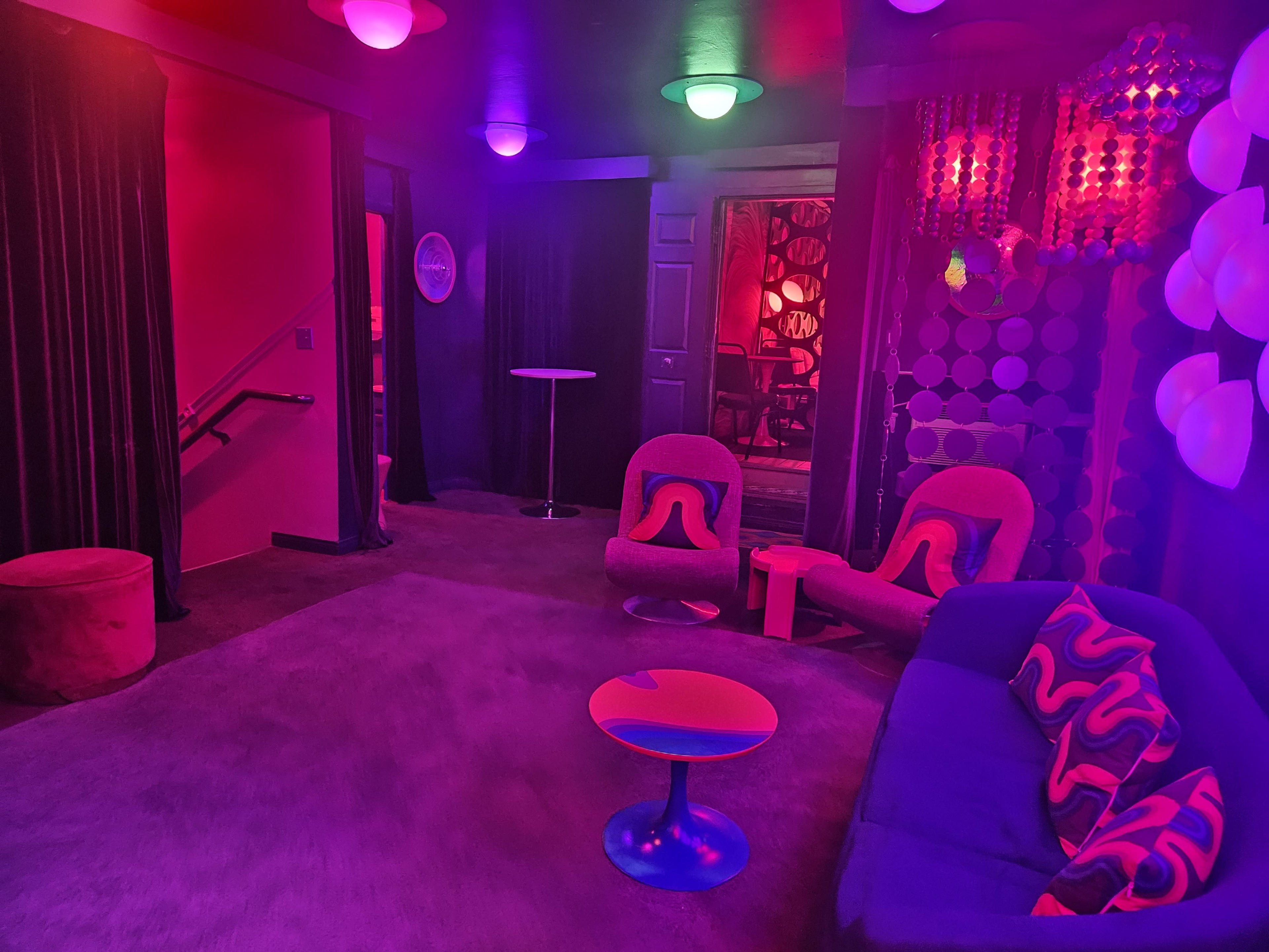 The room features a lounge area with purple and green lighting, soft seating, and round tables surrounded by curtains and patterned walls.