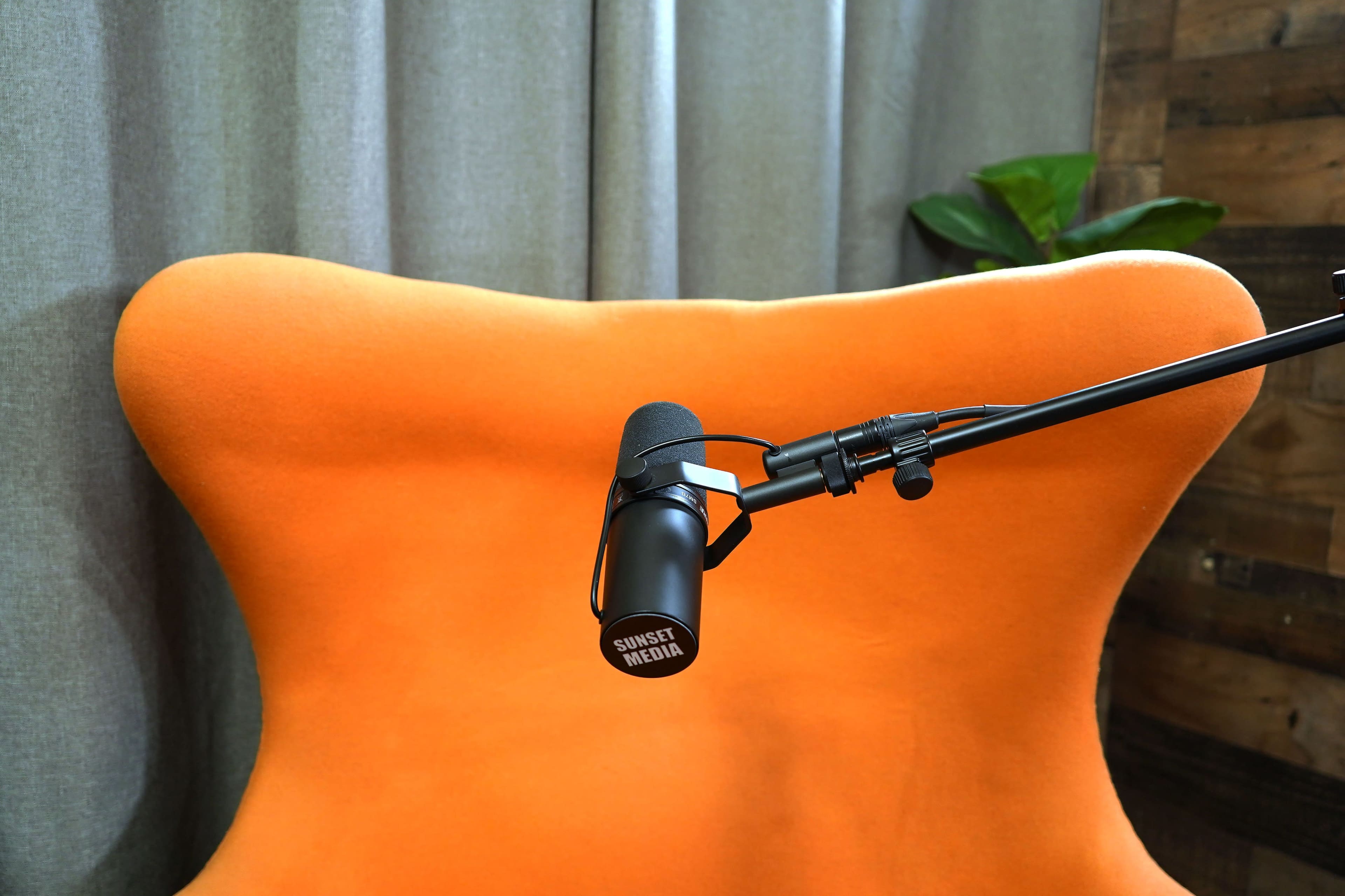 The image shows a bright orange chair with a microphone mounted on an adjustable arm in front of it, set against a backdrop of gray curtains and wooden paneling.