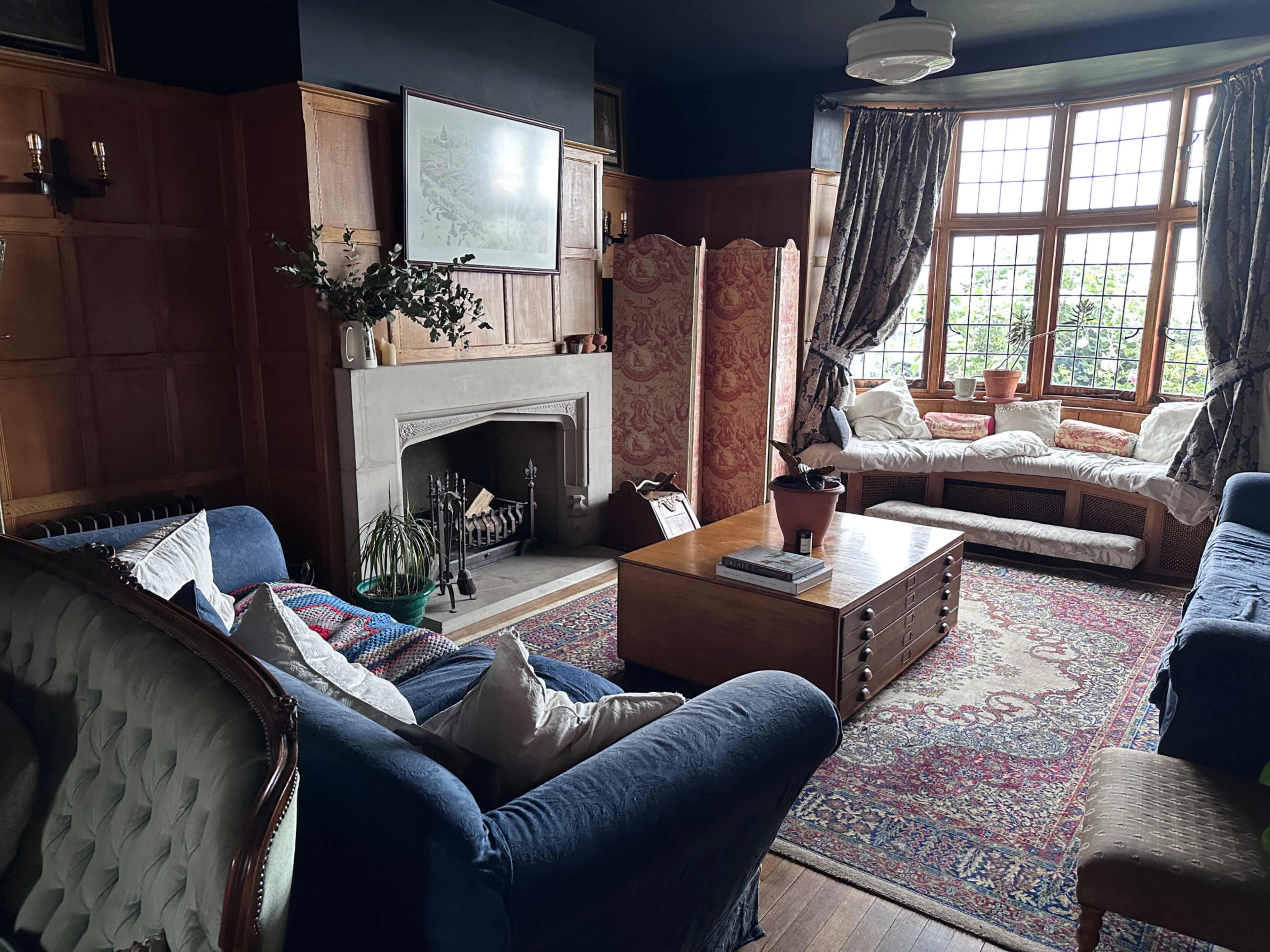 The image shows a cozy living room with wooden paneling, a fireplace, two blue couches, a wooden coffee table, and large windows that let in natural light.