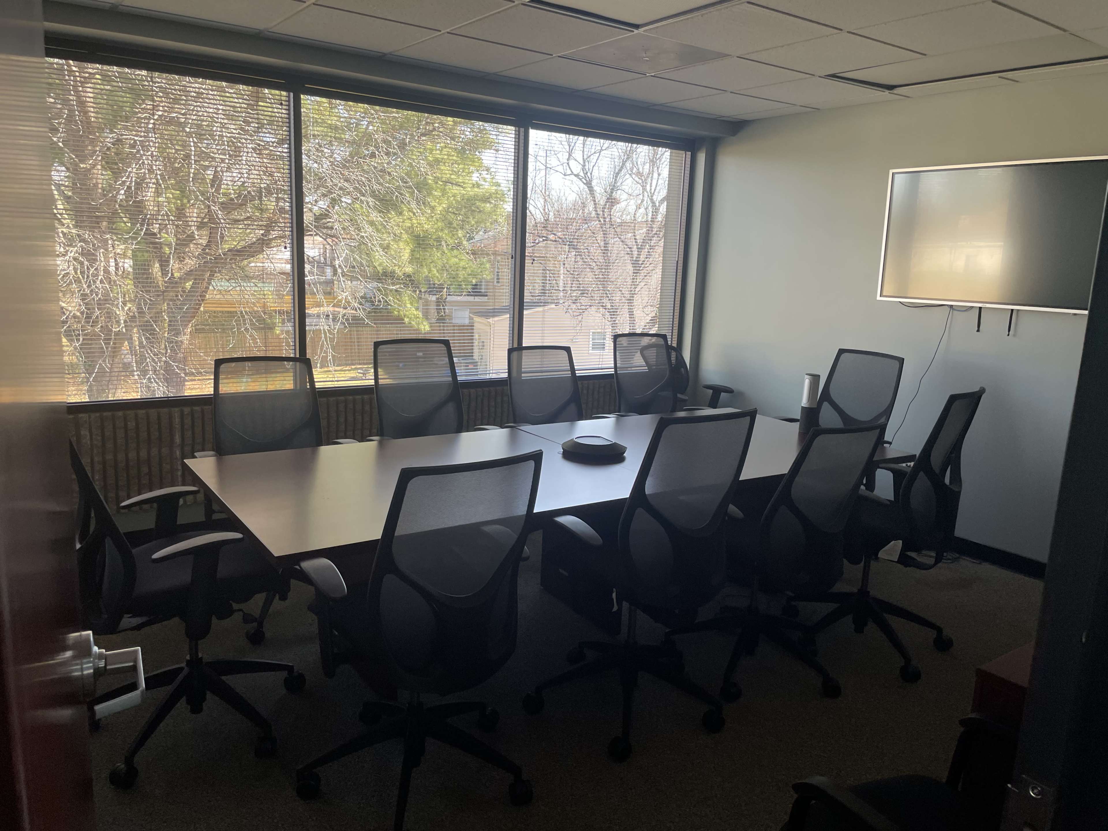 A conference room features a long table surrounded by several rolling chairs and a wall-mounted screen, with large windows providing a view of trees outside.
