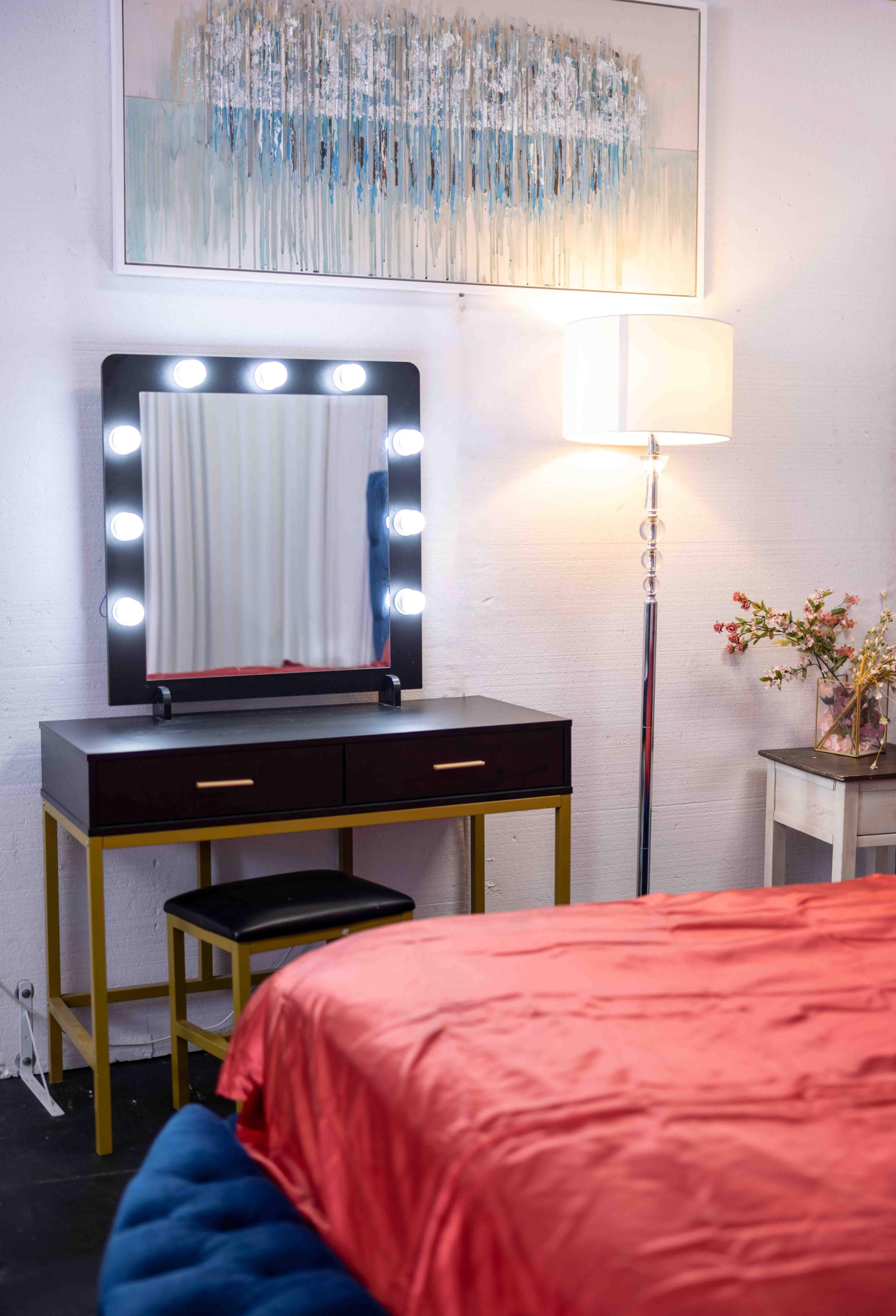 A vanity table with a large illuminated mirror, a lamp, and a bed covered with an orange comforter are arranged in a brightly lit room.
