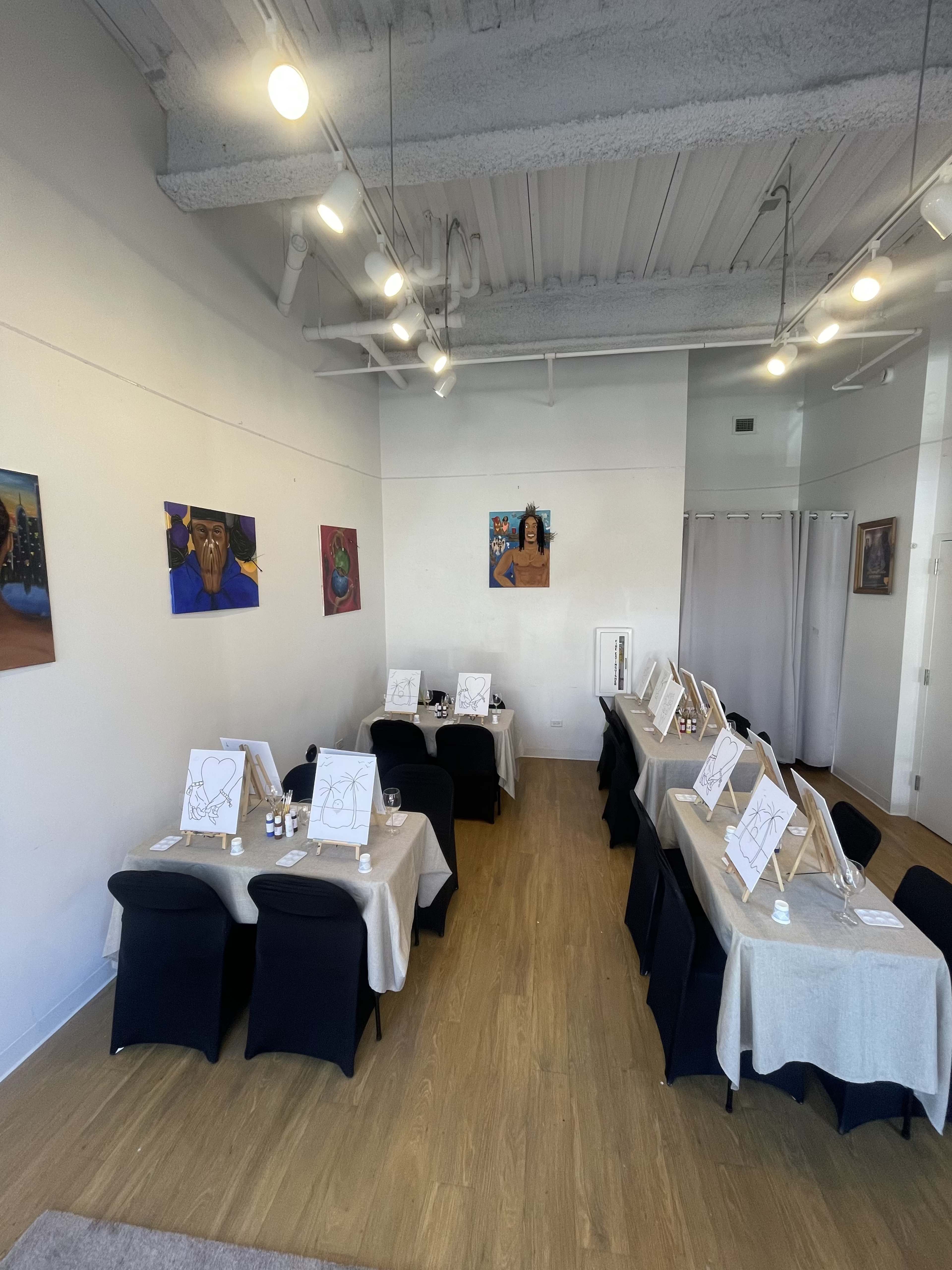 The image shows an art studio with several tables set for a painting session, each with easels, canvases, and paint supplies, along with colorful artwork displayed on the walls.