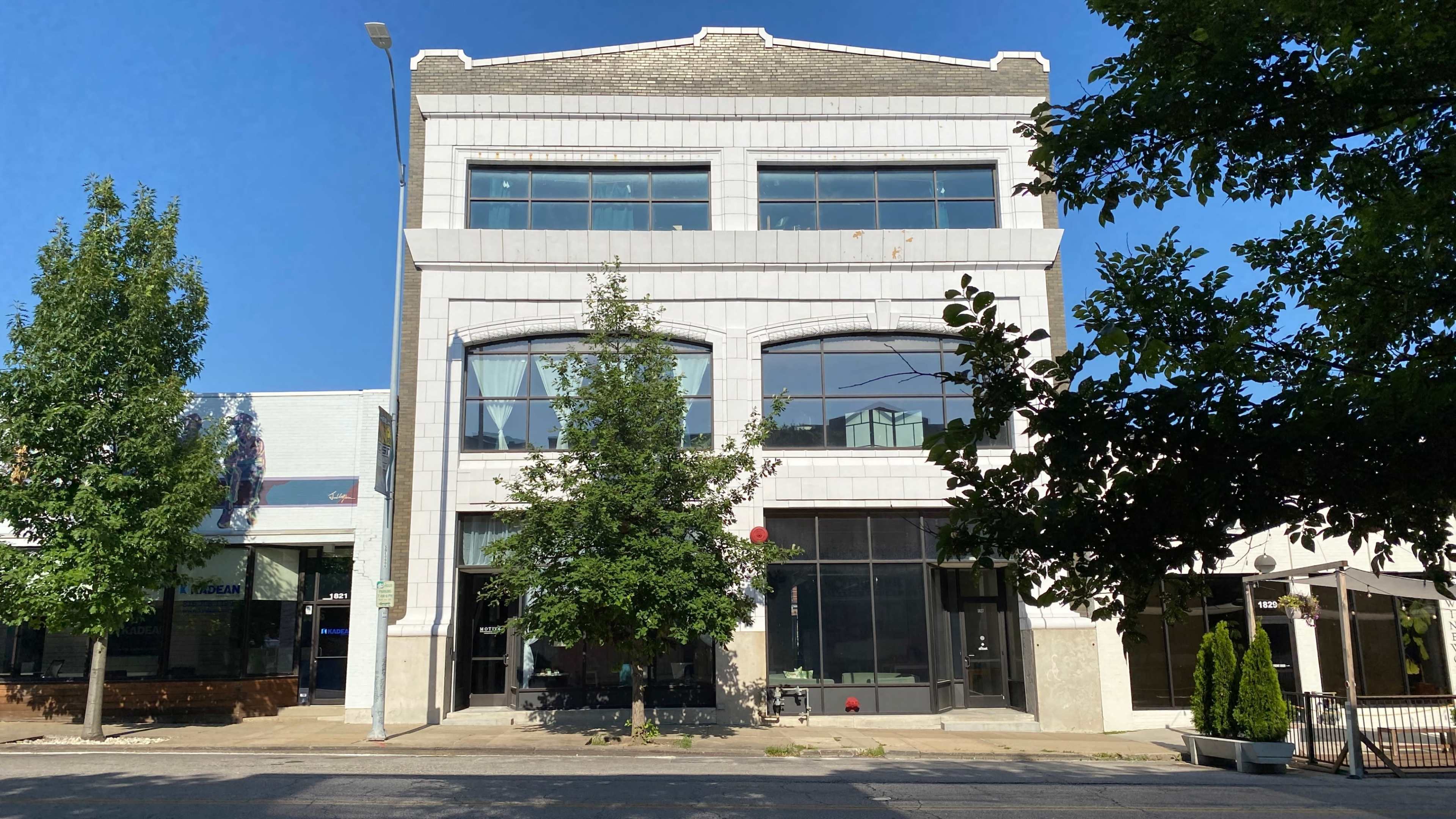 A three-story building with a modern facade and large windows, flanked by smaller commercial structures.