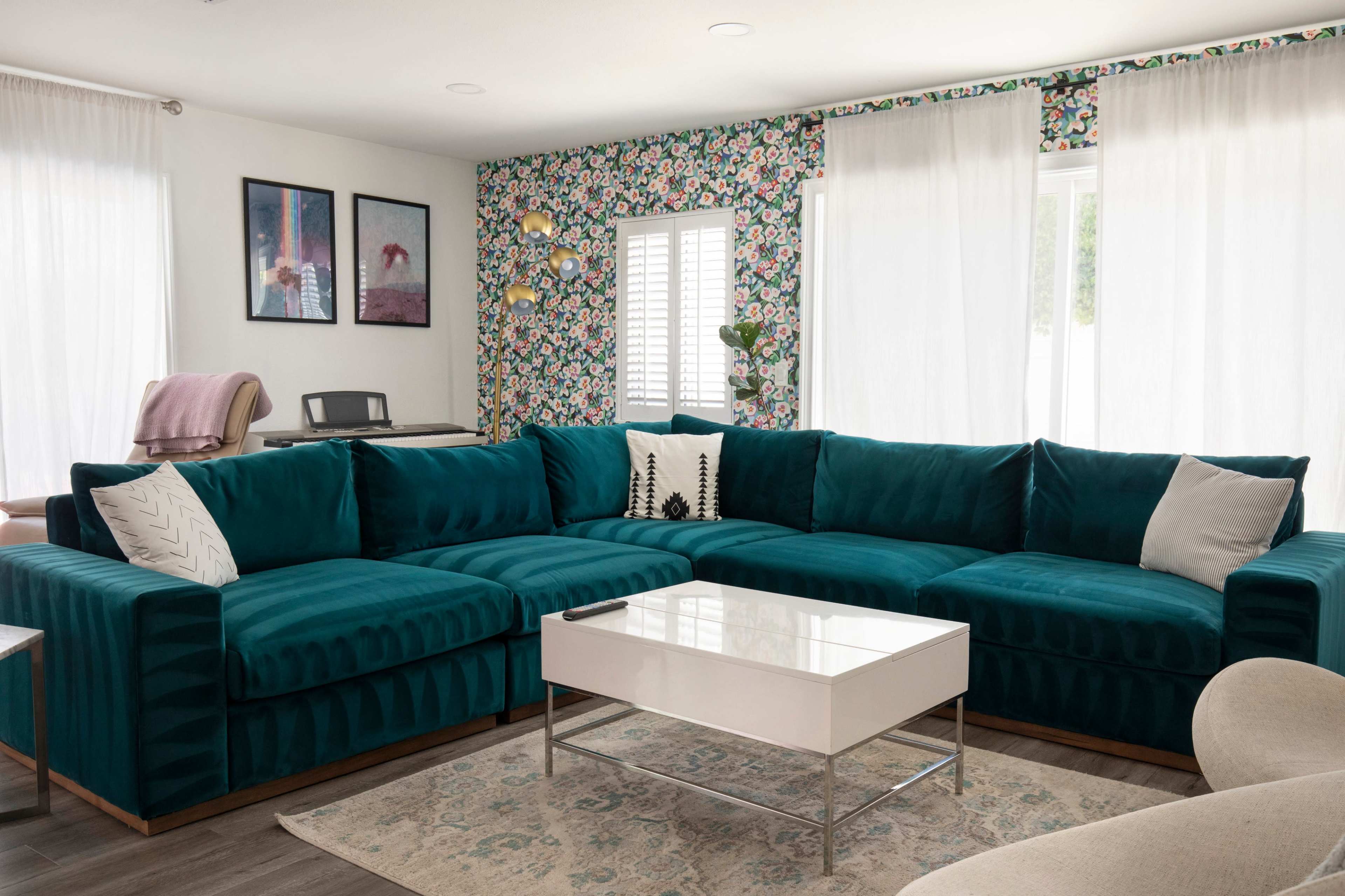 The living room features a large teal sectional sofa, a glass coffee table on a patterned rug, and floral wallpaper adorned with framed artwork.