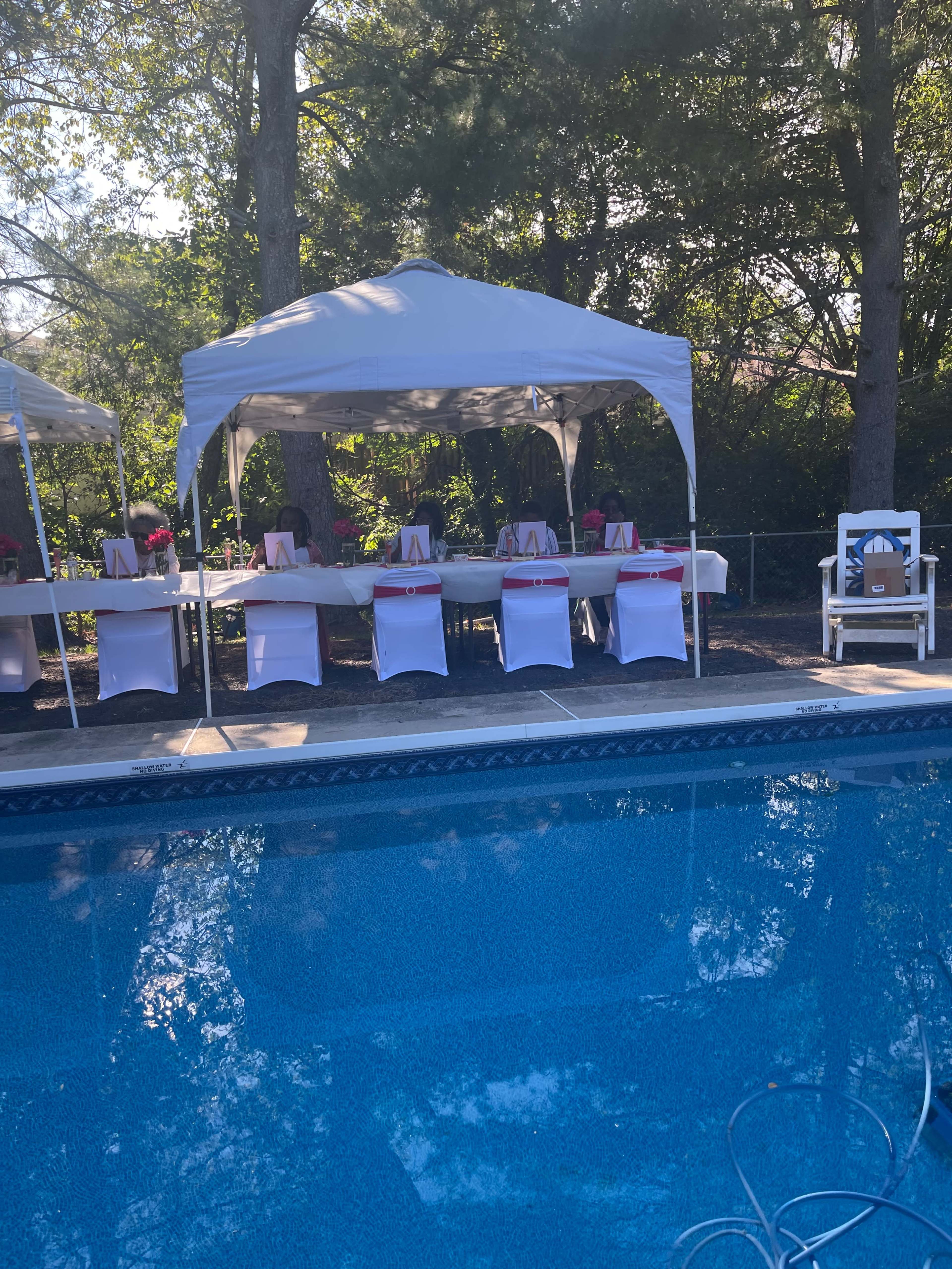 A poolside area features several white tents with tables set up, adorned with red accents and chairs, surrounded by trees.
