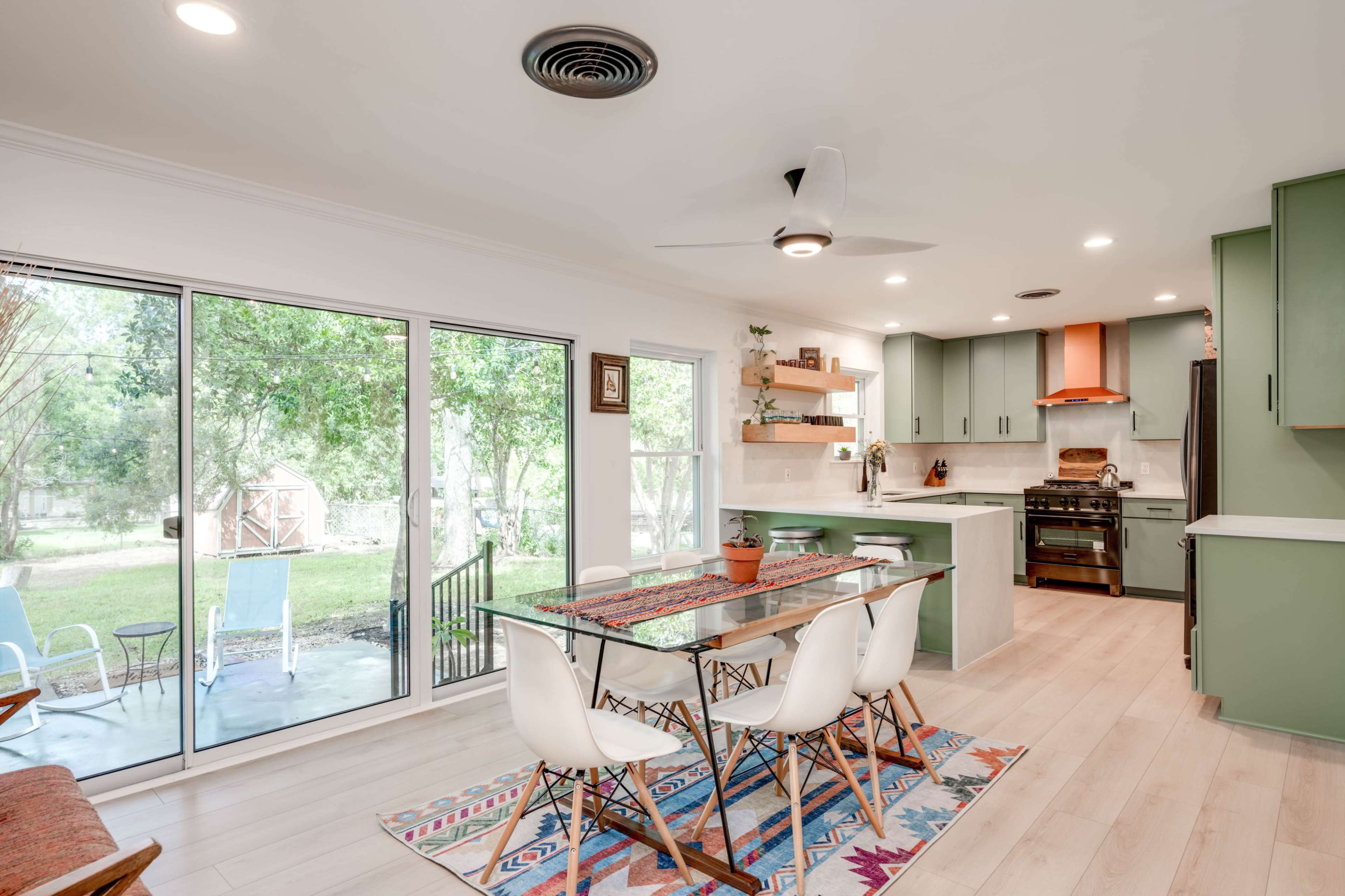 A modern kitchen and dining area features a glass table surrounded by white chairs, with large windows offering a view of the backyard.