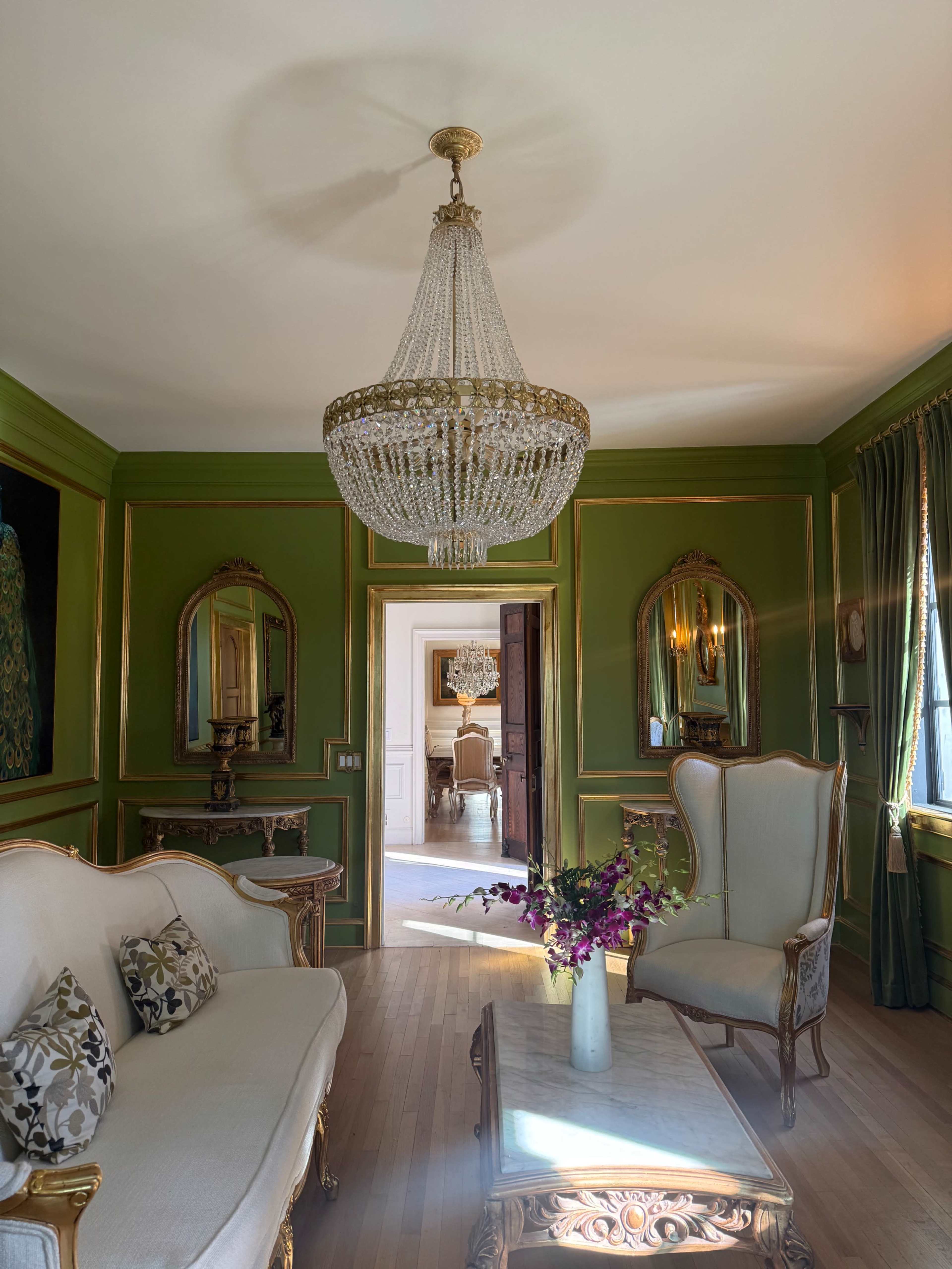 The room features a crystal chandelier, green walls with gold accents, and elegant furniture, including a sofa and armchair, with a view of a dining area in the background.