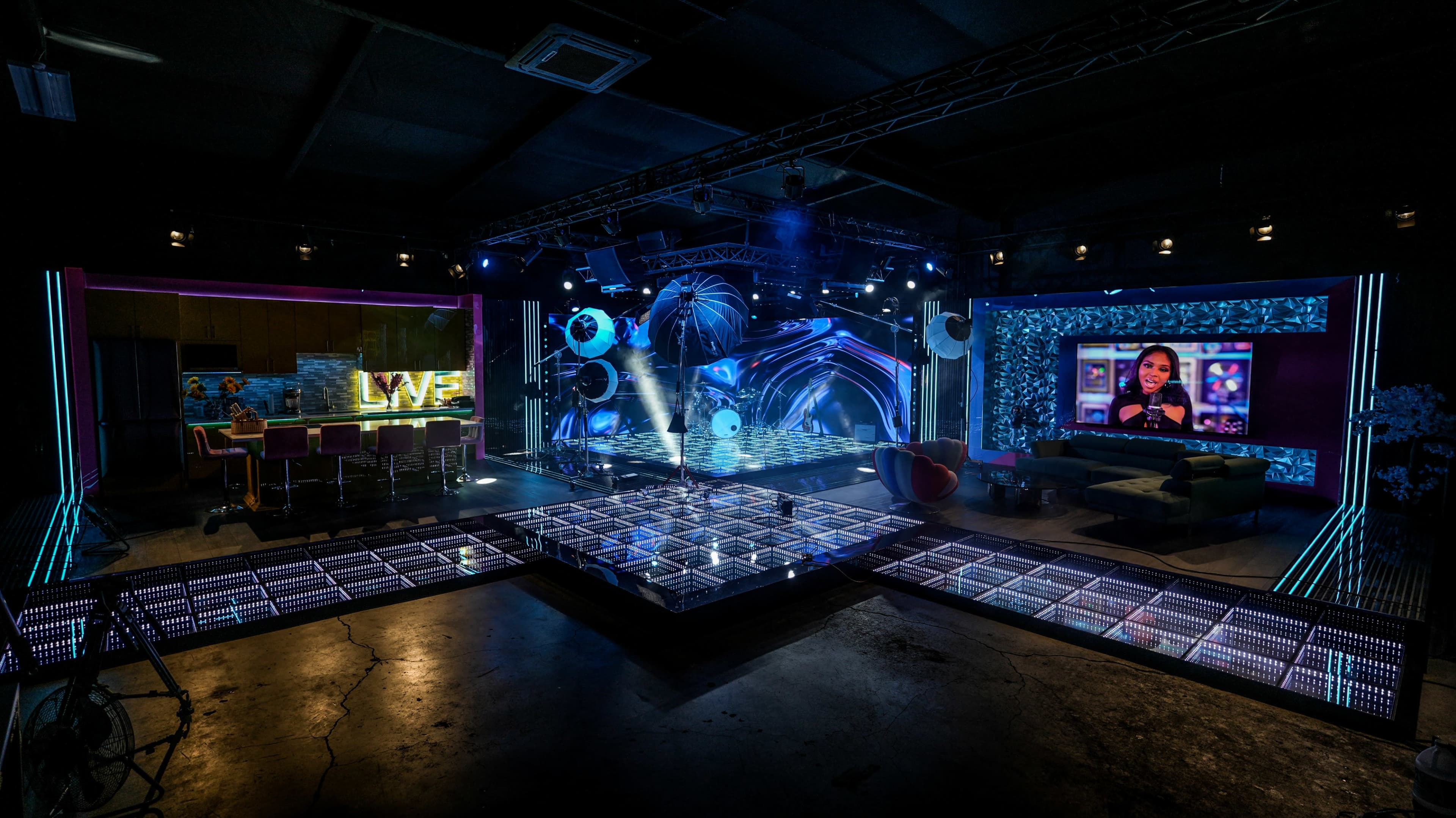 The image shows a modern television studio set with colorful lighting, a large screen displaying a host, and a glass-paneled floor area with seating arrangements.