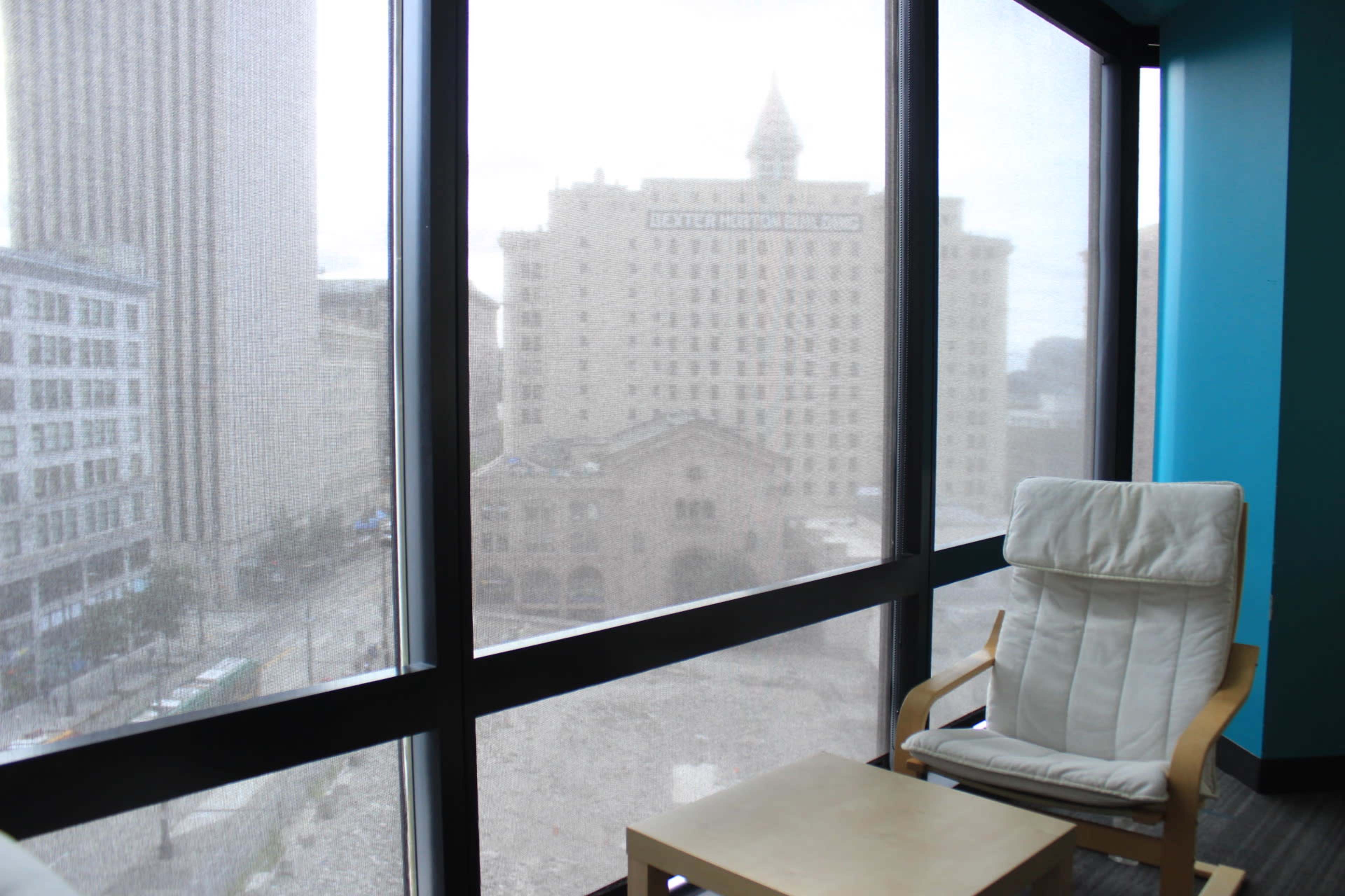 A chair and small table are positioned by a large window overlooking a city street and buildings in the background.