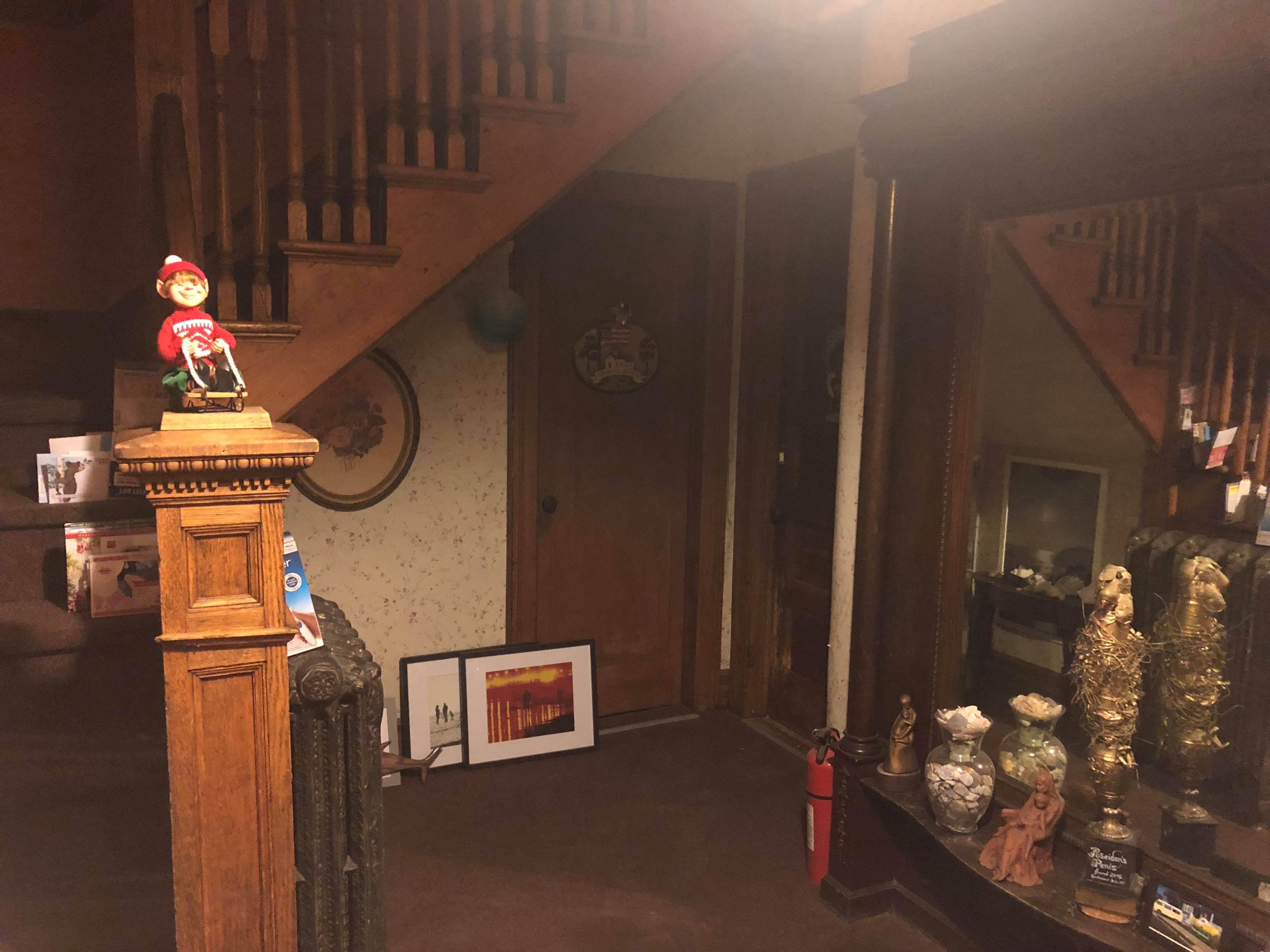The image shows a dimly lit entrance area with a staircase, a wooden banister, and various decorative items displayed along the walls and on furniture.