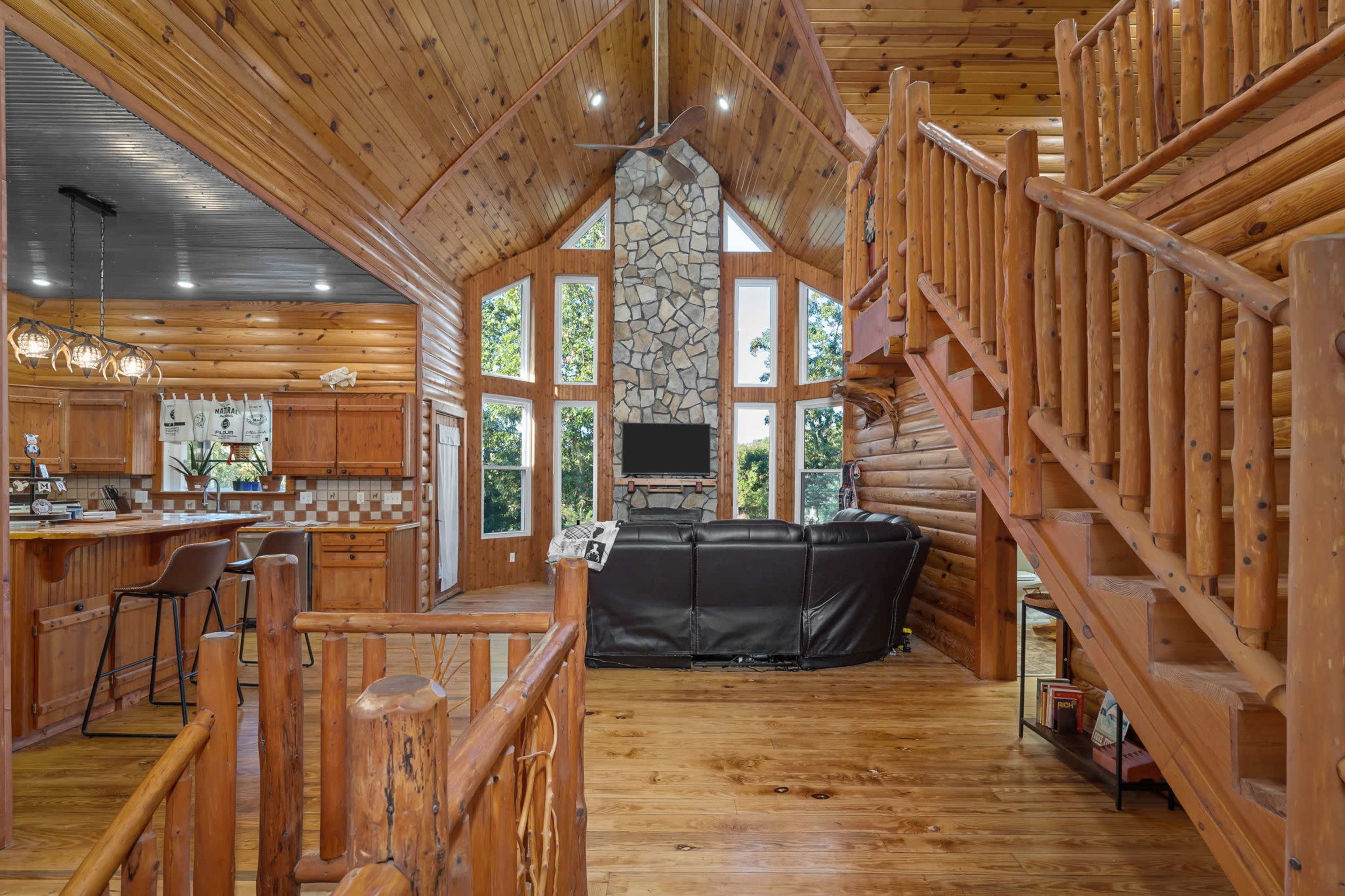 A wooden interior of a log cabin featuring a spacious living area with a stone fireplace, large windows, and a staircase leading to an upper level.