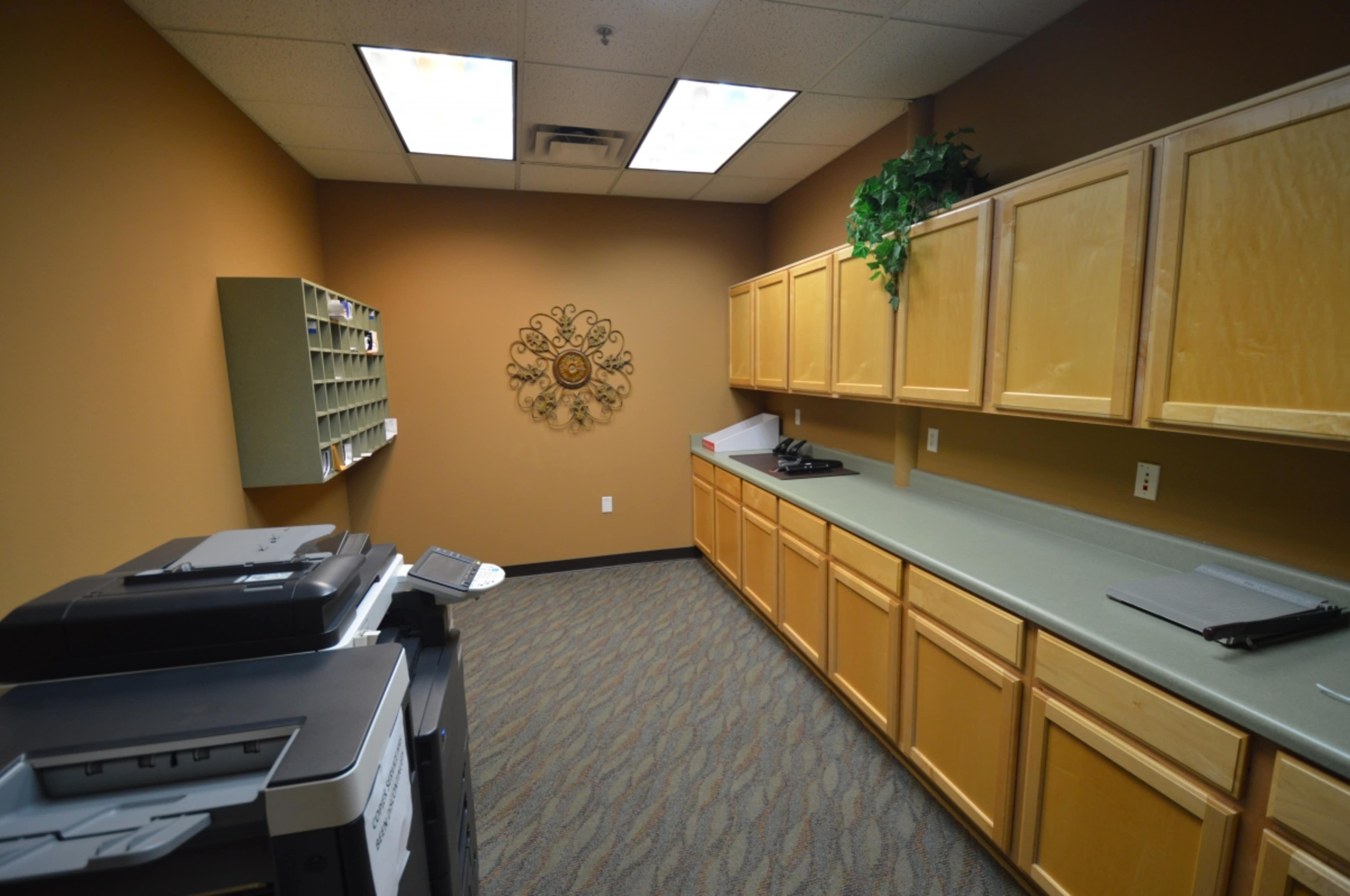 A small office break room with wooden cabinets, a countertop, a printer, and a wall-mounted shelf for mail or documents.