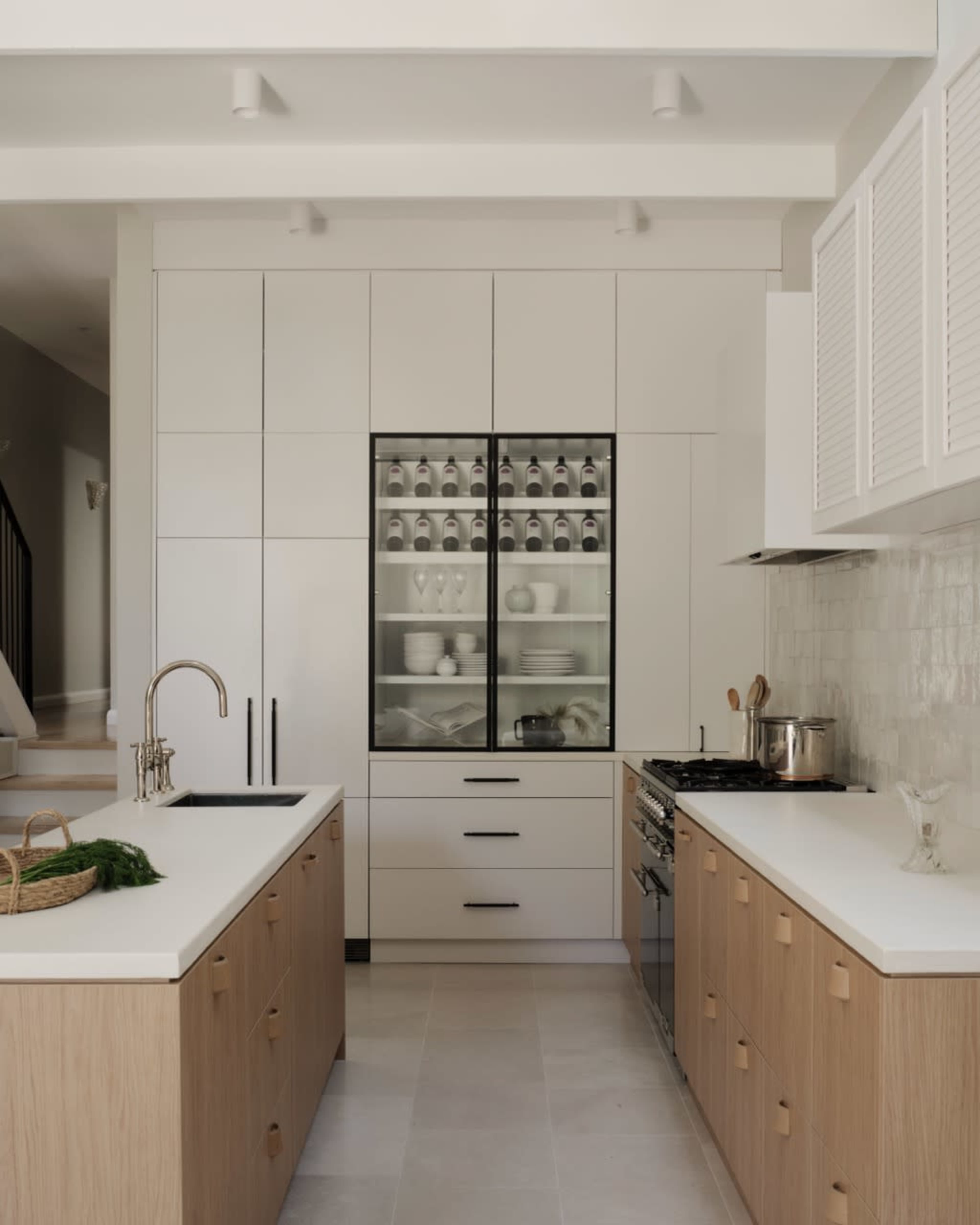 The image shows a modern kitchen with light cabinetry, a glass-fronted cabinet displaying dinnerware, and a central island with a sink and stove.