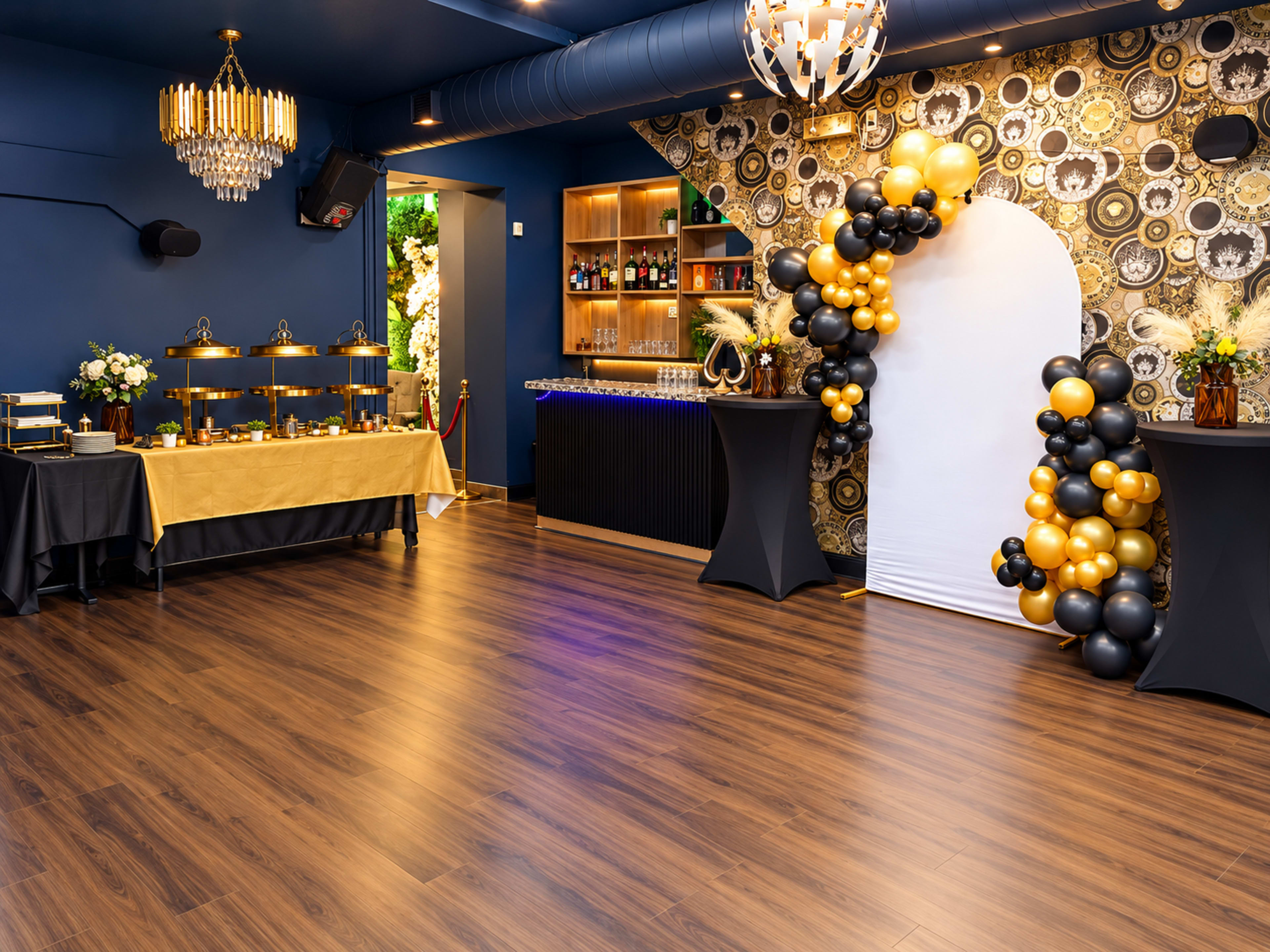 The image shows a decorated event space featuring a bar, a buffet table, and a balloon arch in black and gold against a patterned wall.