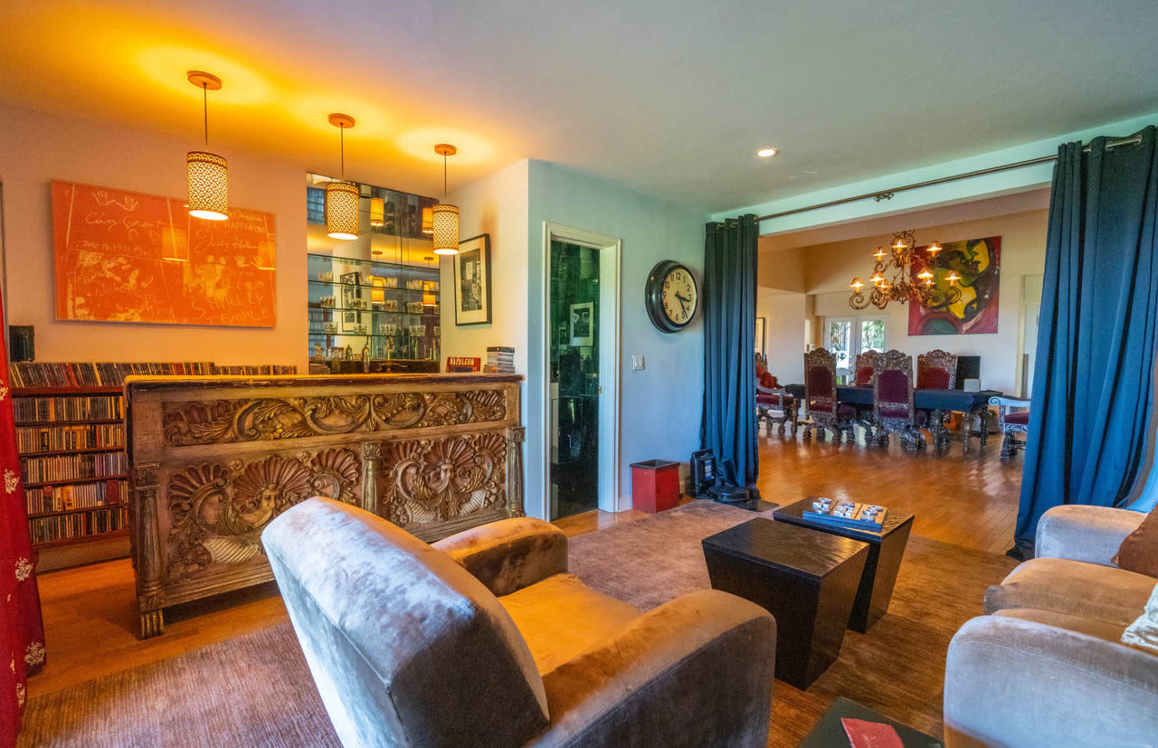 The image shows a spacious living area featuring a decorative bar, lounge seating, and a view of a separate room with elaborate chairs and a chandelier.