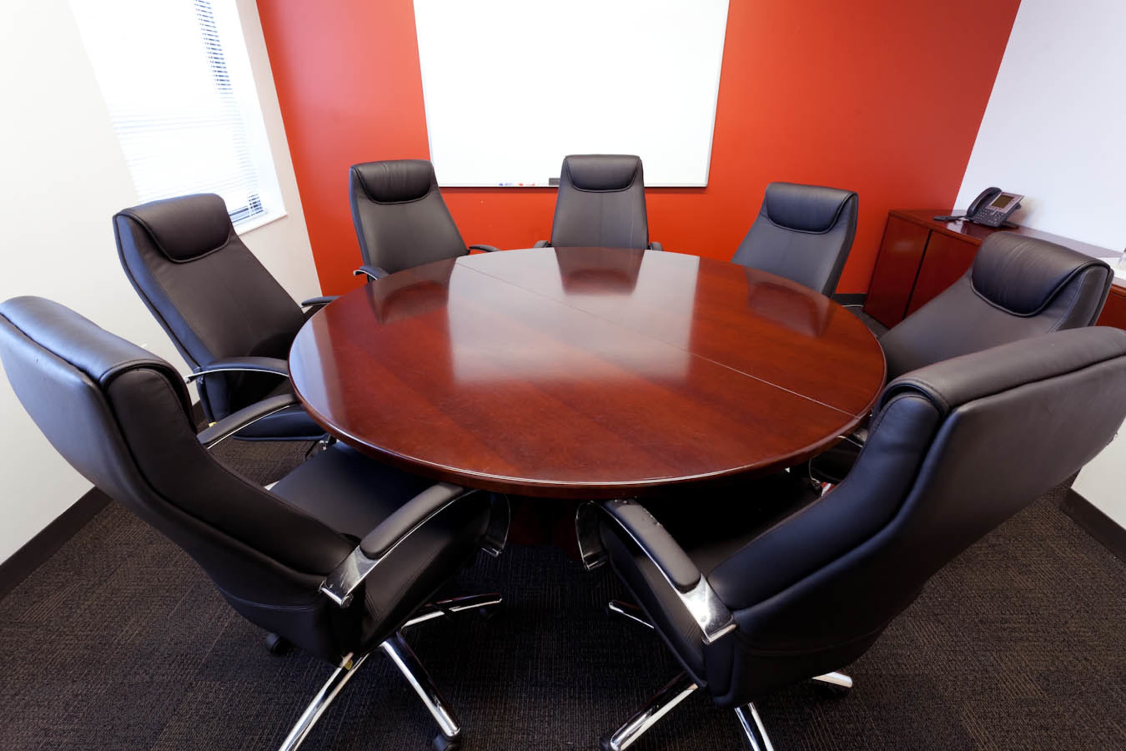 A round wooden conference table is set in a meeting room with six black leather chairs arranged around it.
