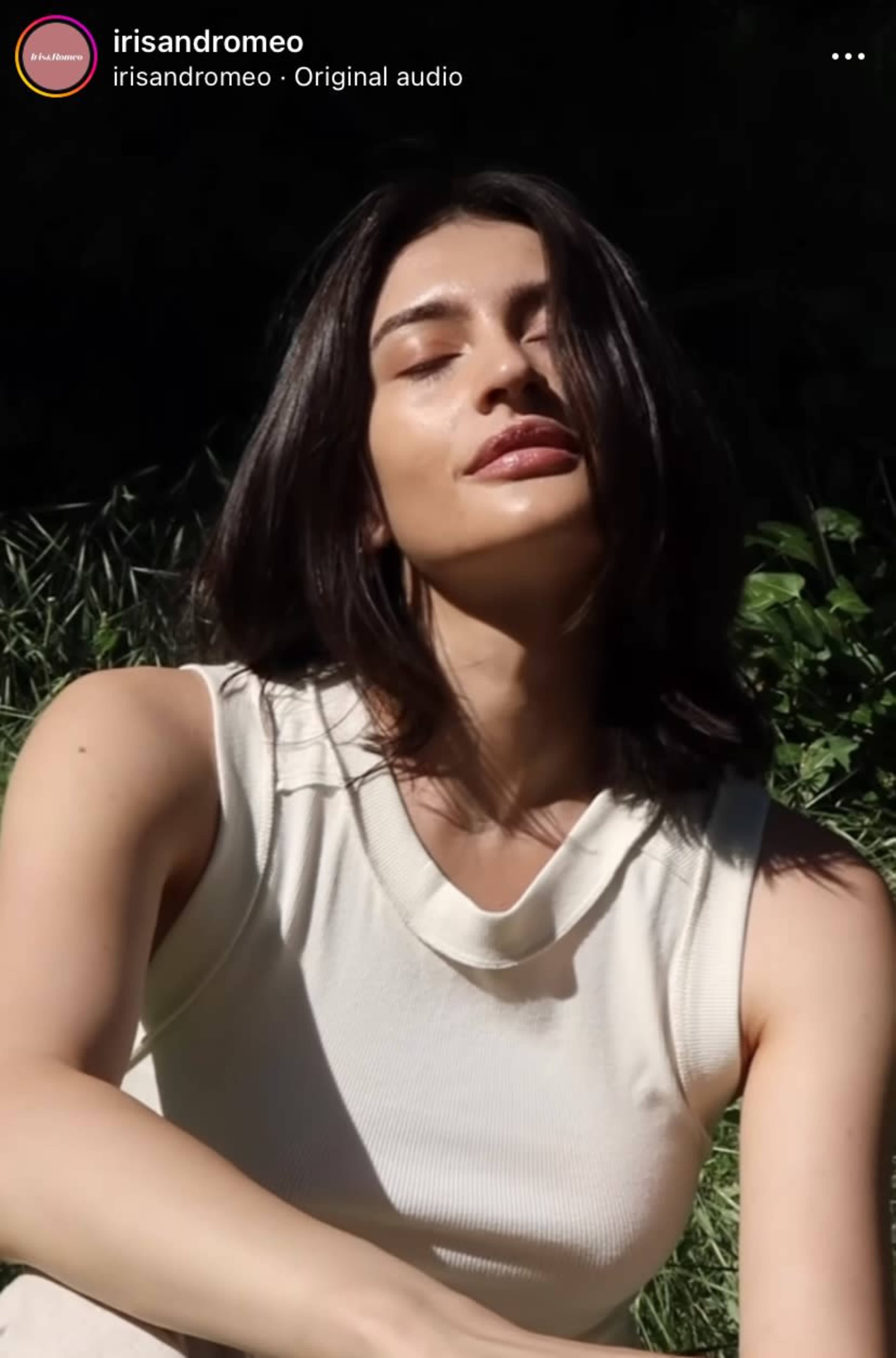 A person with long hair is sitting outdoors in a grassy area, wearing a sleeveless, light-colored top.