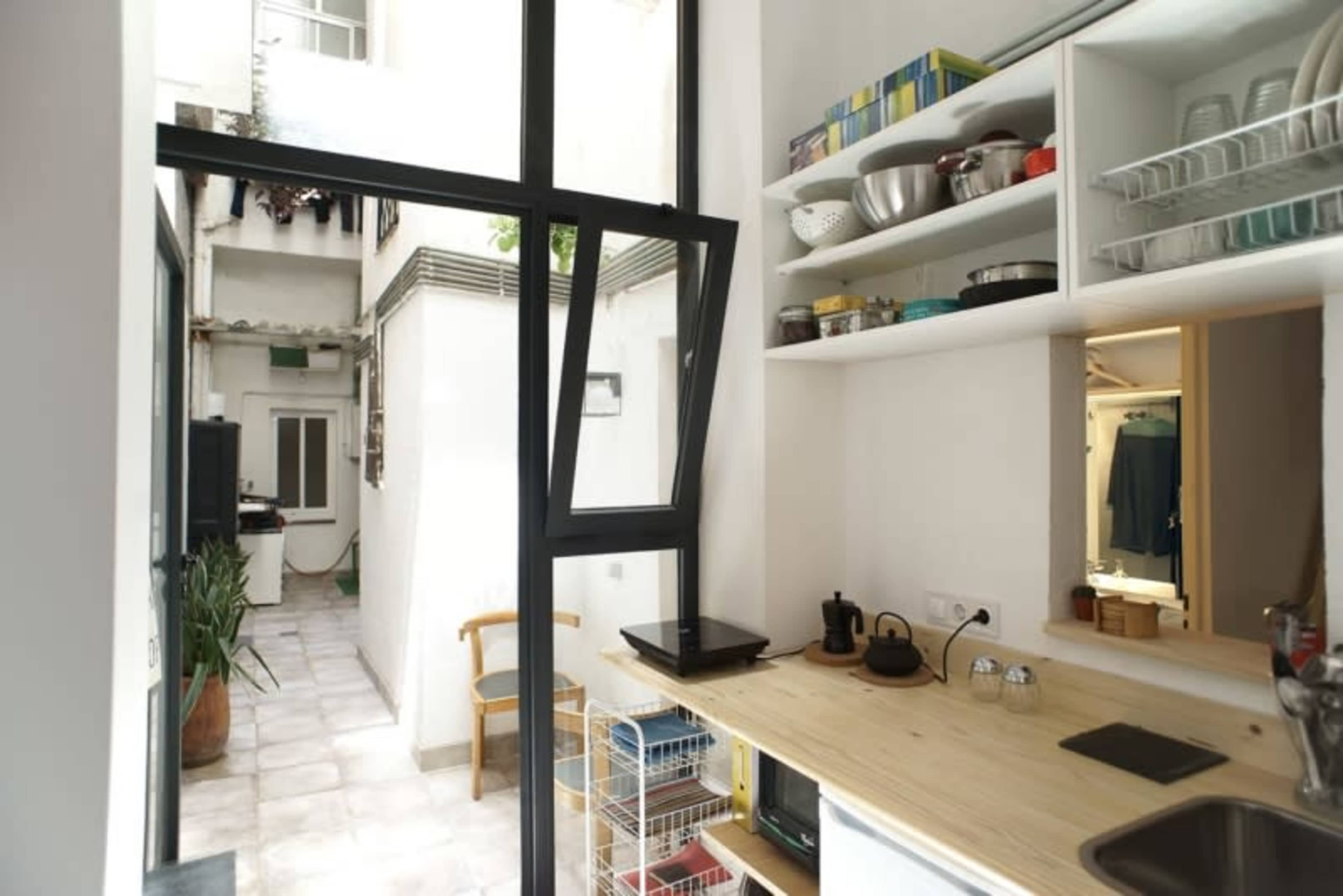 A small kitchen with a wooden countertop, shelves of dishware, and an open window overlooking a narrow courtyard.