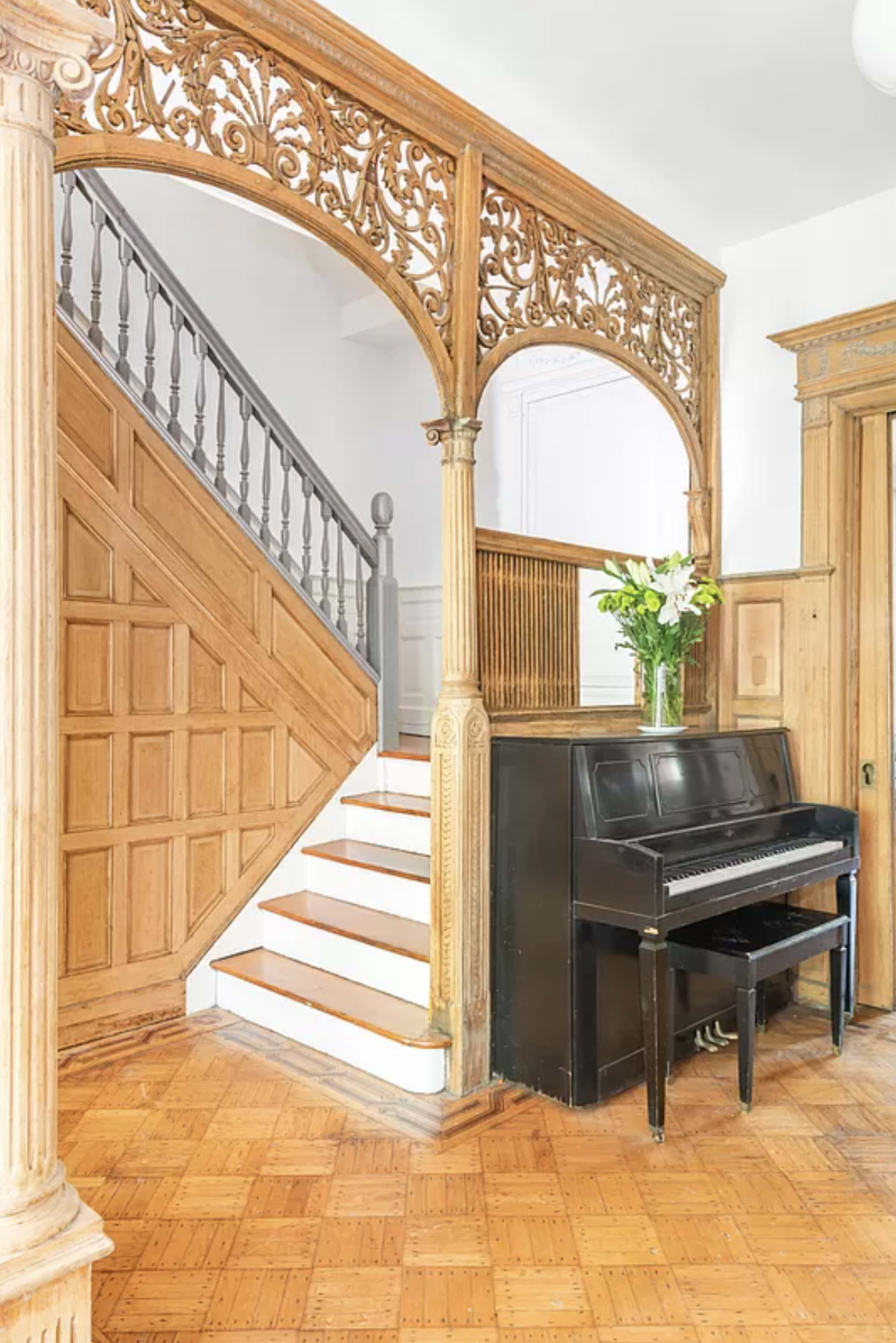 The image depicts an elegant entryway featuring a wooden staircase, decorative wooden archway, and a black piano positioned against the wall.